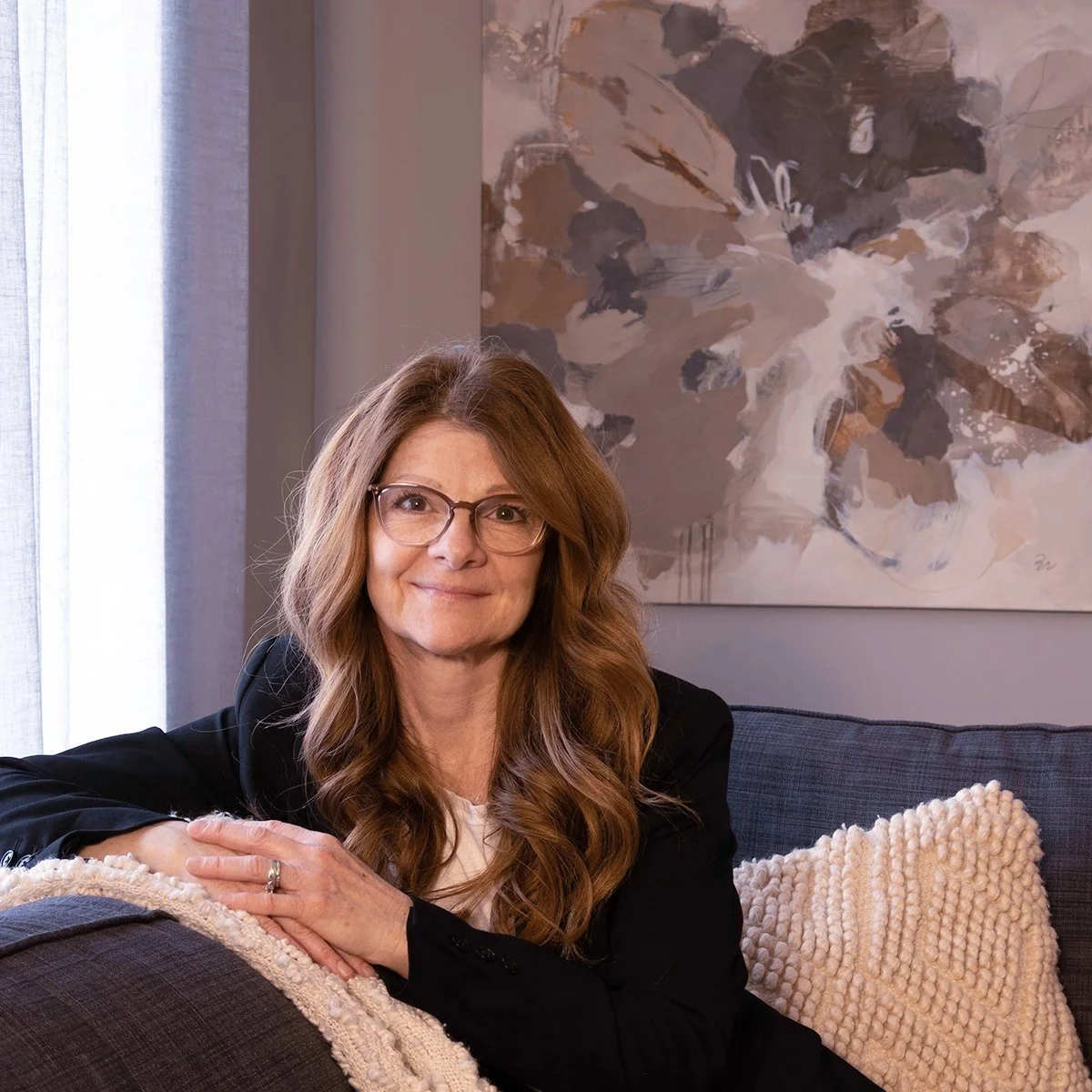 A woman with long, wavy red hair and glasses, wearing a black jacket, sitting on a sofa with a cream-colored textured pillow, in a room with abstract wall art and a sheer curtain.