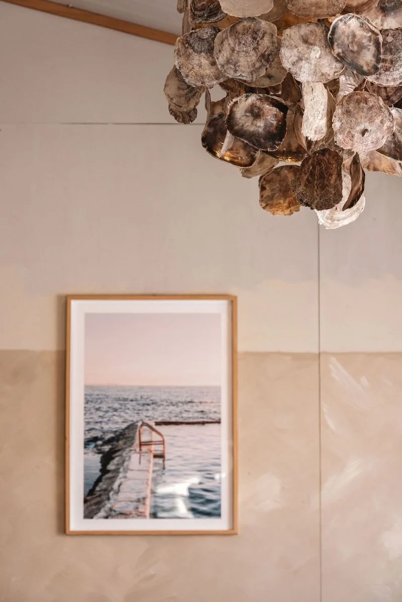 A framed photograph of a beach scene with a pier extending into the water, hanging on a beige wall, with a Oyster chandelier  hanging above.