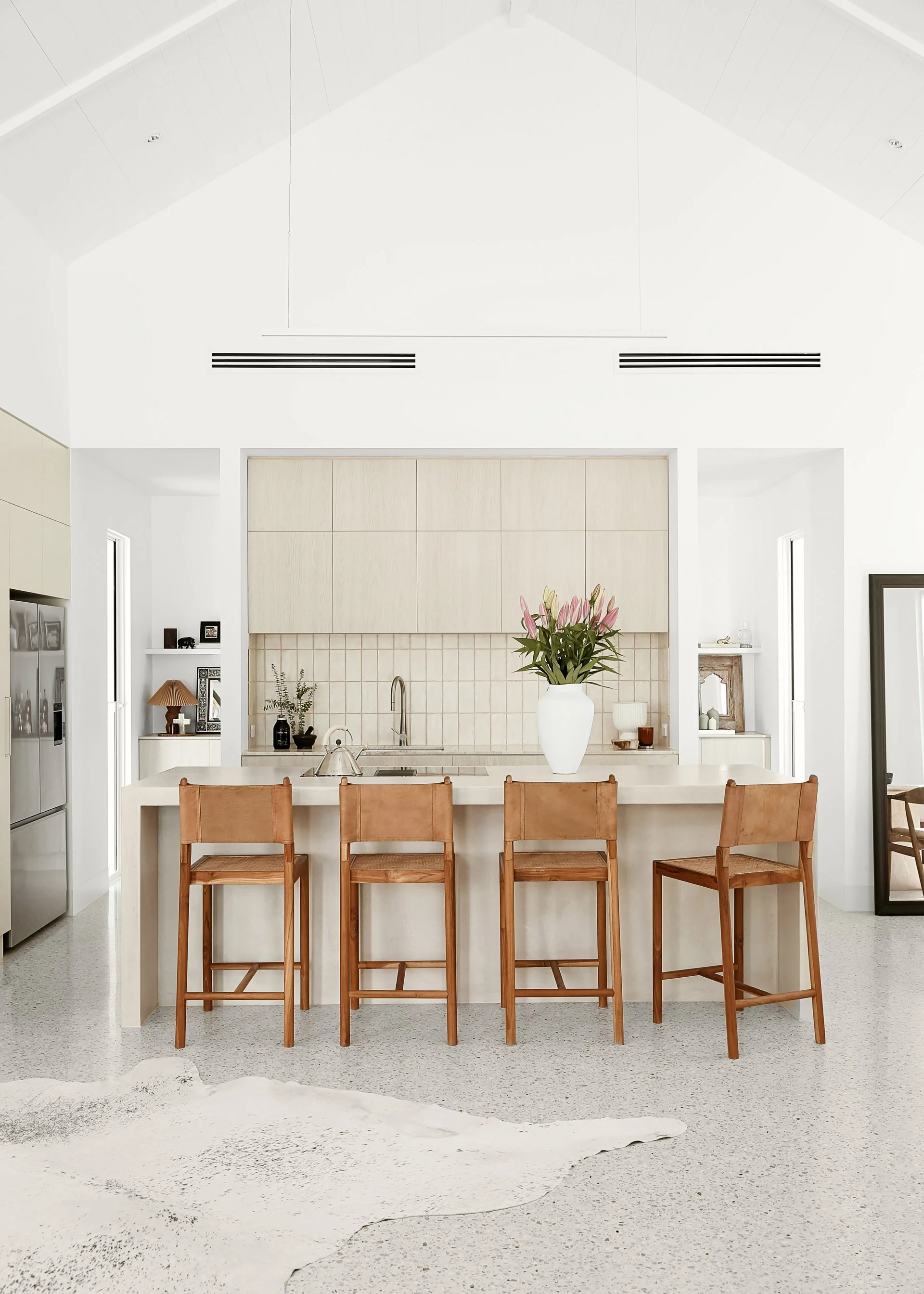 Modern kitchen and dining area with a white kitchen island, four wooden chairs, a large white vase with pink flowers, and built-in cabinets.