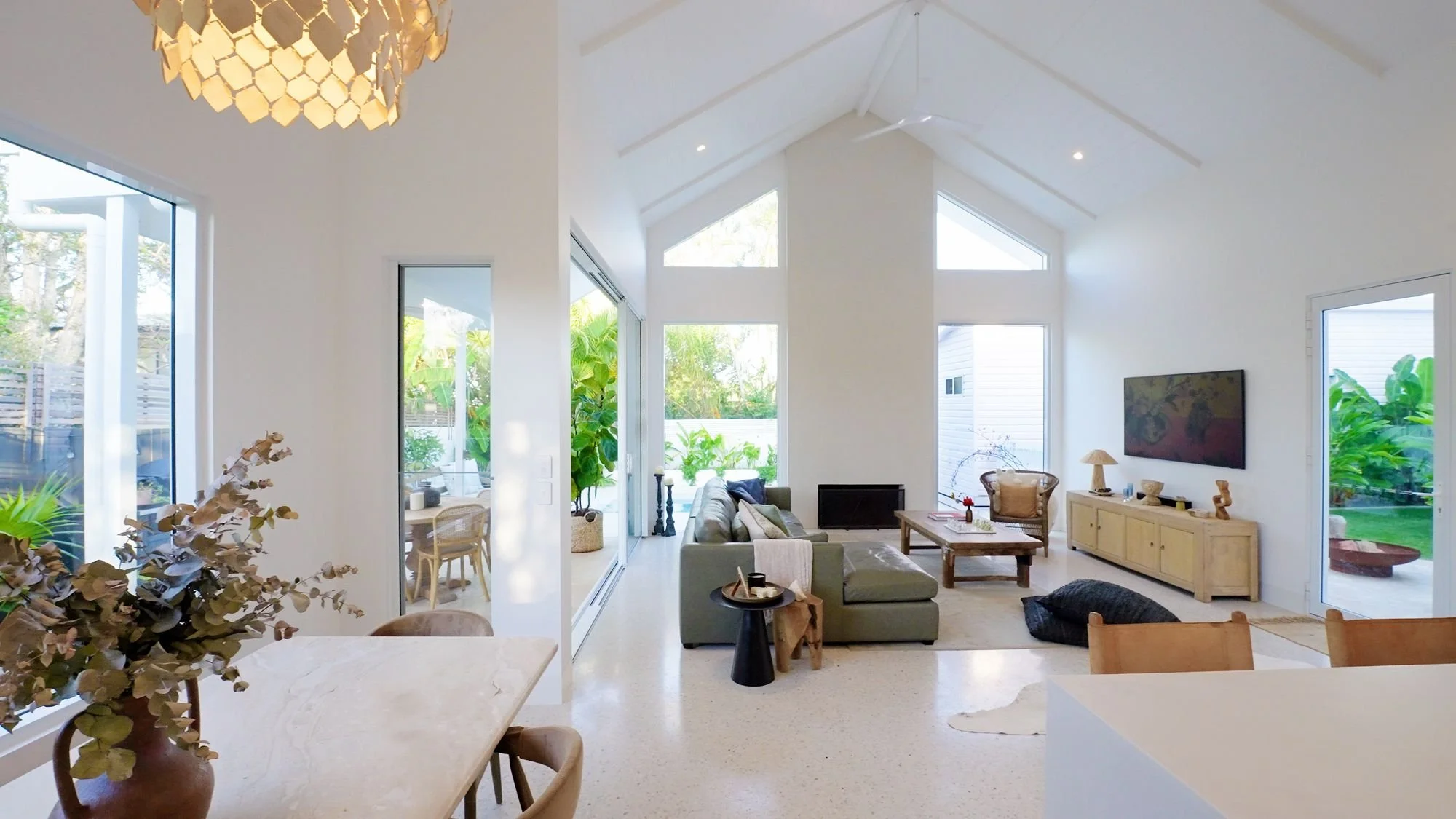 Living room with large windows, white walls, vaulted ceiling, and modern furniture including a green sofa, a wooden coffee table, a beige TV console, and a wall-mounted large flat-screen TV. There is a ceiling fan, plants, and decorative items throug