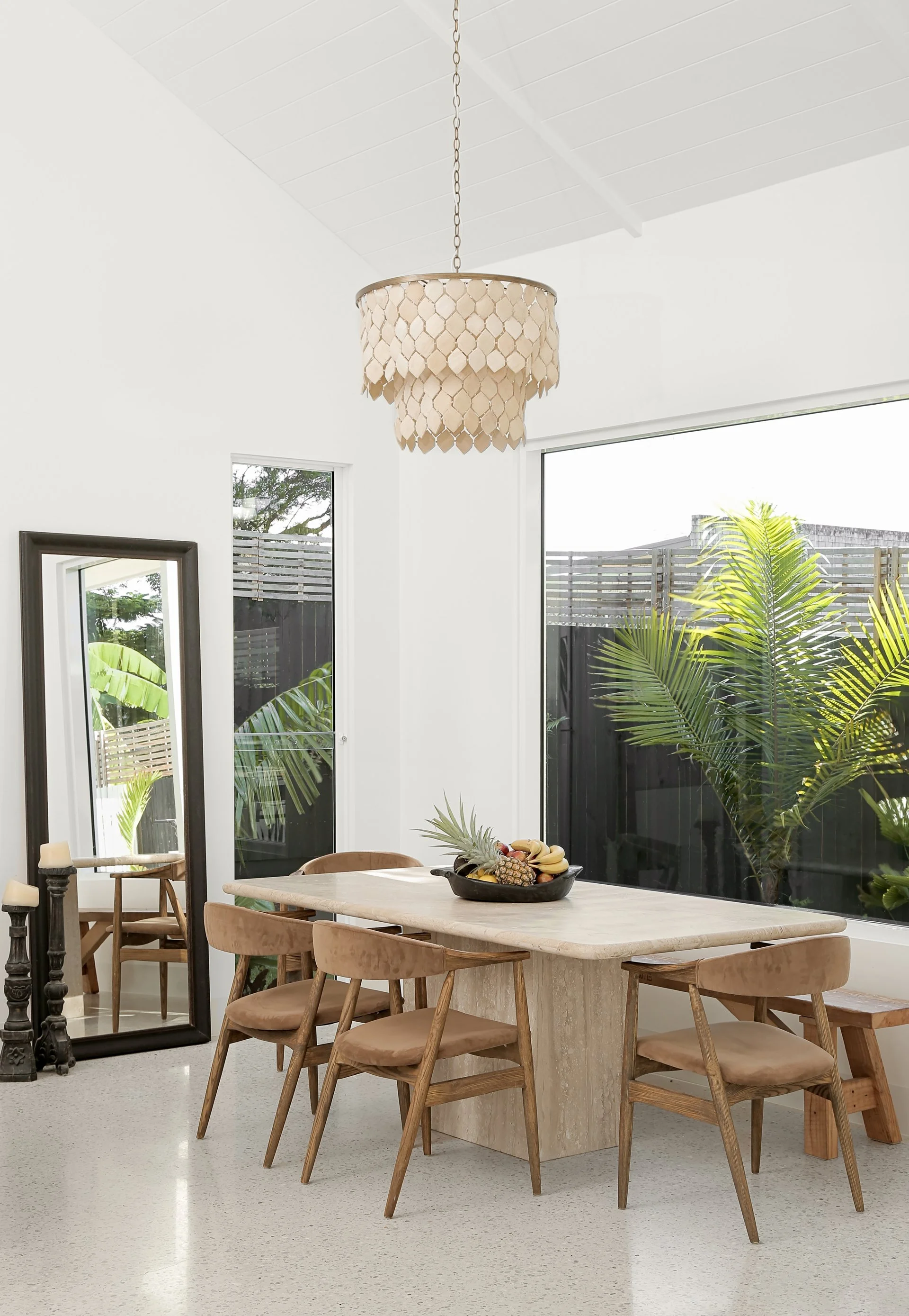 A dining room with a light-colored stone table, six wooden chairs with tan cushions, a black fruit bowl with bananas and pineapple, large windows with plants outside, a mirror, and a hanging beige lamp with a scalloped design.