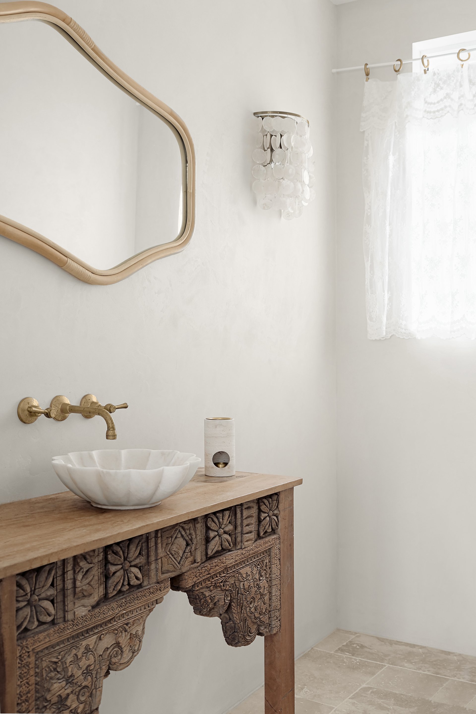 A bathroom with a carved wooden vanity, a white ceramic vessel sink, a brass faucet, a mirror, a wall light fixture with shell decorations, and a window with lace curtains.