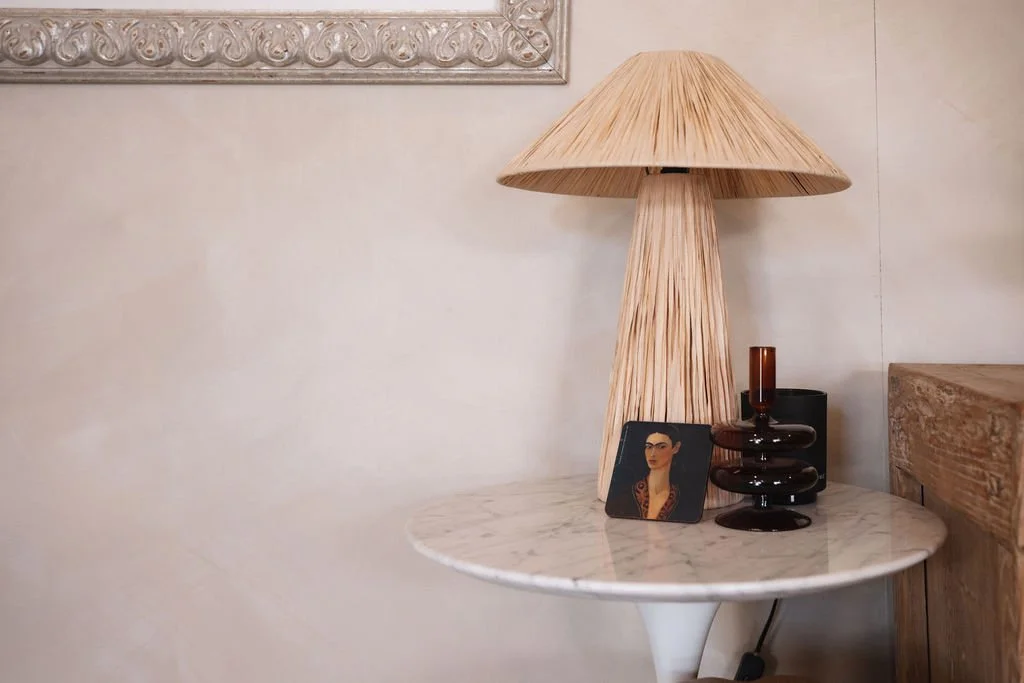 A round marble side table with a beige textured lamp, a small portrait painting of a woman, and two dark glass vases on it, against a light-colored wall.