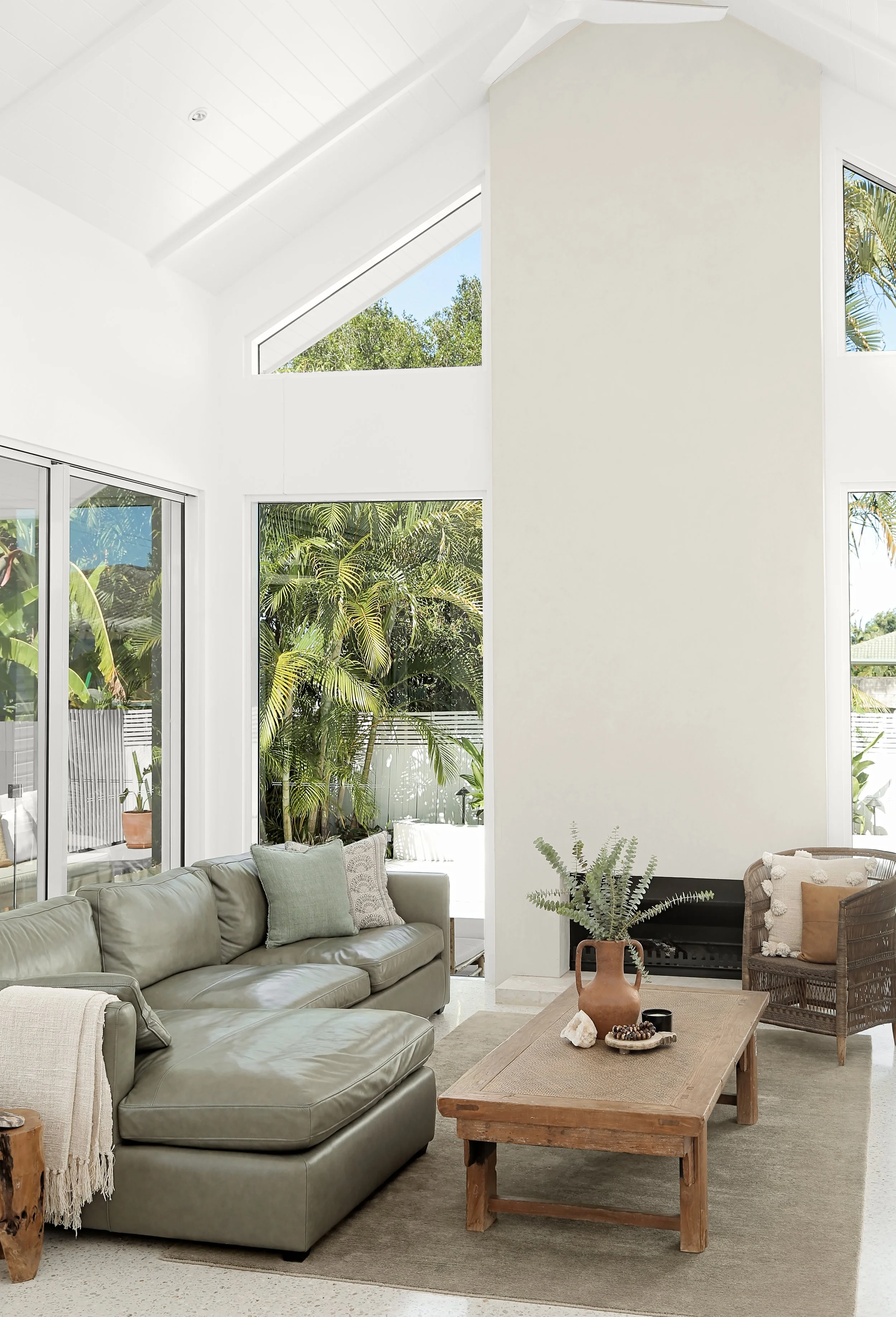A living room with a beige leather sofa (with gray and beige pillows), a wooden coffee table with decorative items including a large vase with a fern, a bowl, and rocks, a woven chair with a beige pillow, large windows revealing green outdoor foliage
