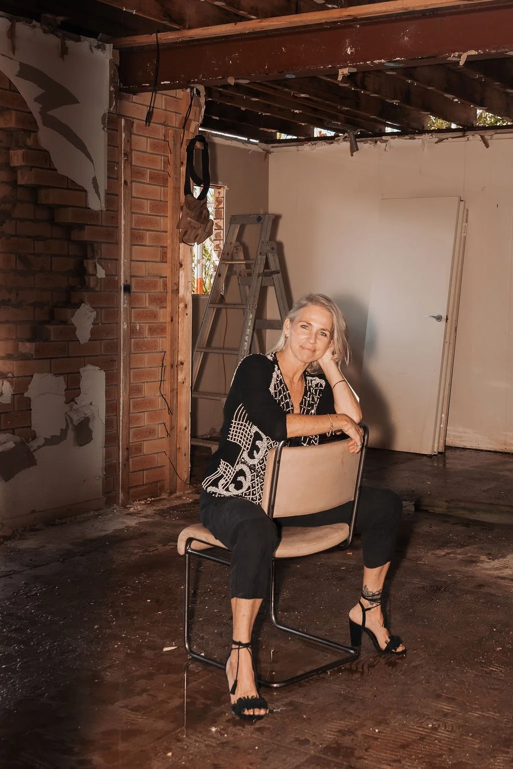 Sarah Wood sitting on a beige chair in a partially renovated room with exposed brick wall, wooden beams, and renovation tools. She is smiling and posing with her head resting on her hand.