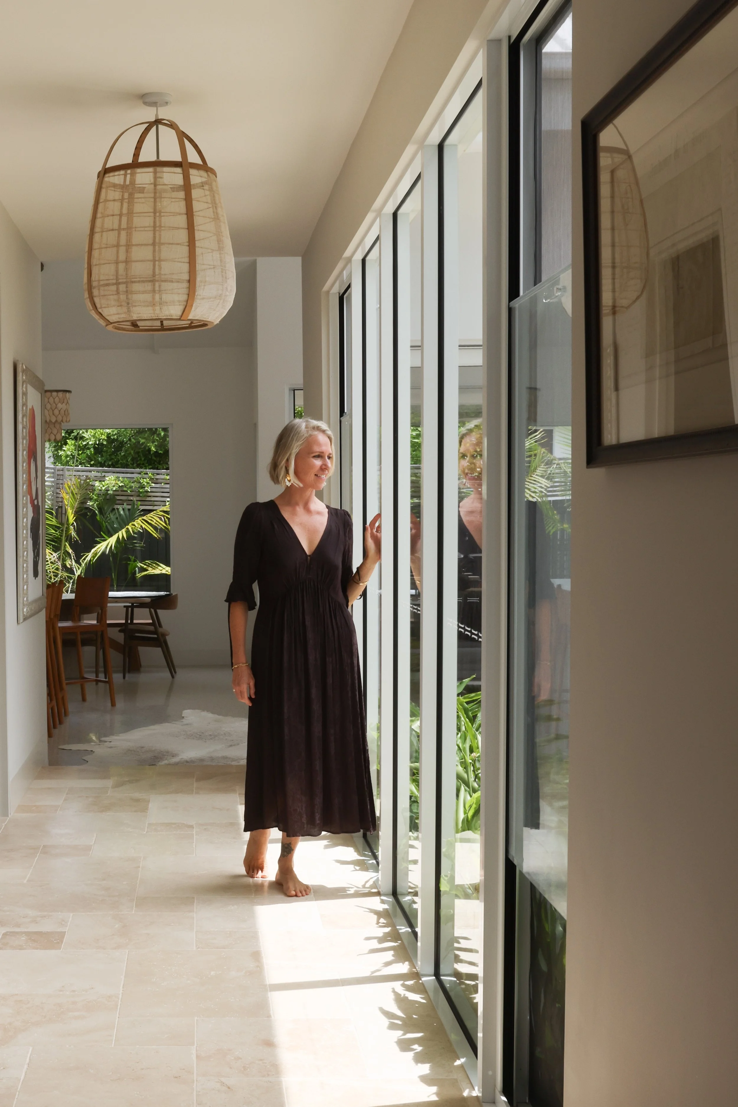 Sarah Wood in a long black dress is walking barefoot through a room with large glass doors, sunlight casting shadows, and greenery outside.  The room is decorated with framed pictures and hanging light fixtures.