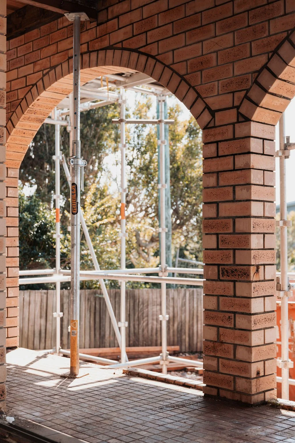 View through a brick archway with scaffolding, construction equipment, and a wooden fence outside.