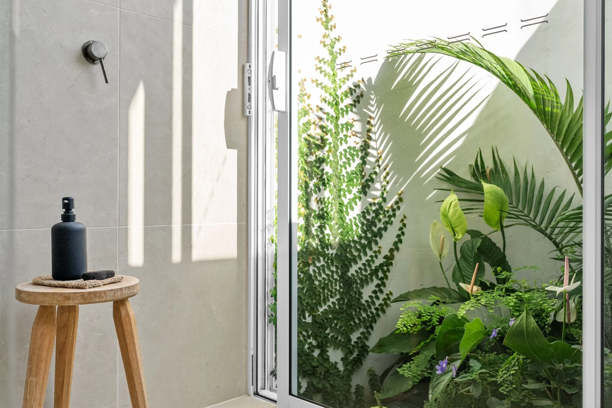 A bathroom with a glass door that opens to a small garden area with green leafy plants and ivy. Inside, a wooden stool holds a black soap dispenser, a sponge, and a small black container. Sunlight casts shadows, creating a bright and natural atmosphe