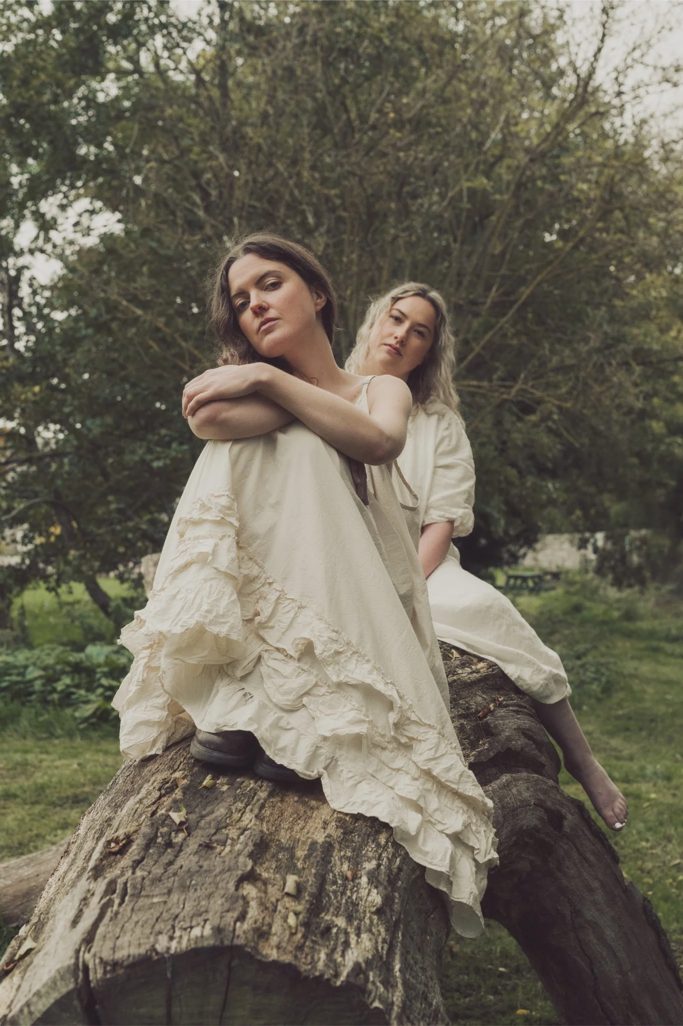 Two women dressed in cream-colored, vintage-style dresses sitting on a large fallen tree in a lush green outdoor setting with trees in the background.