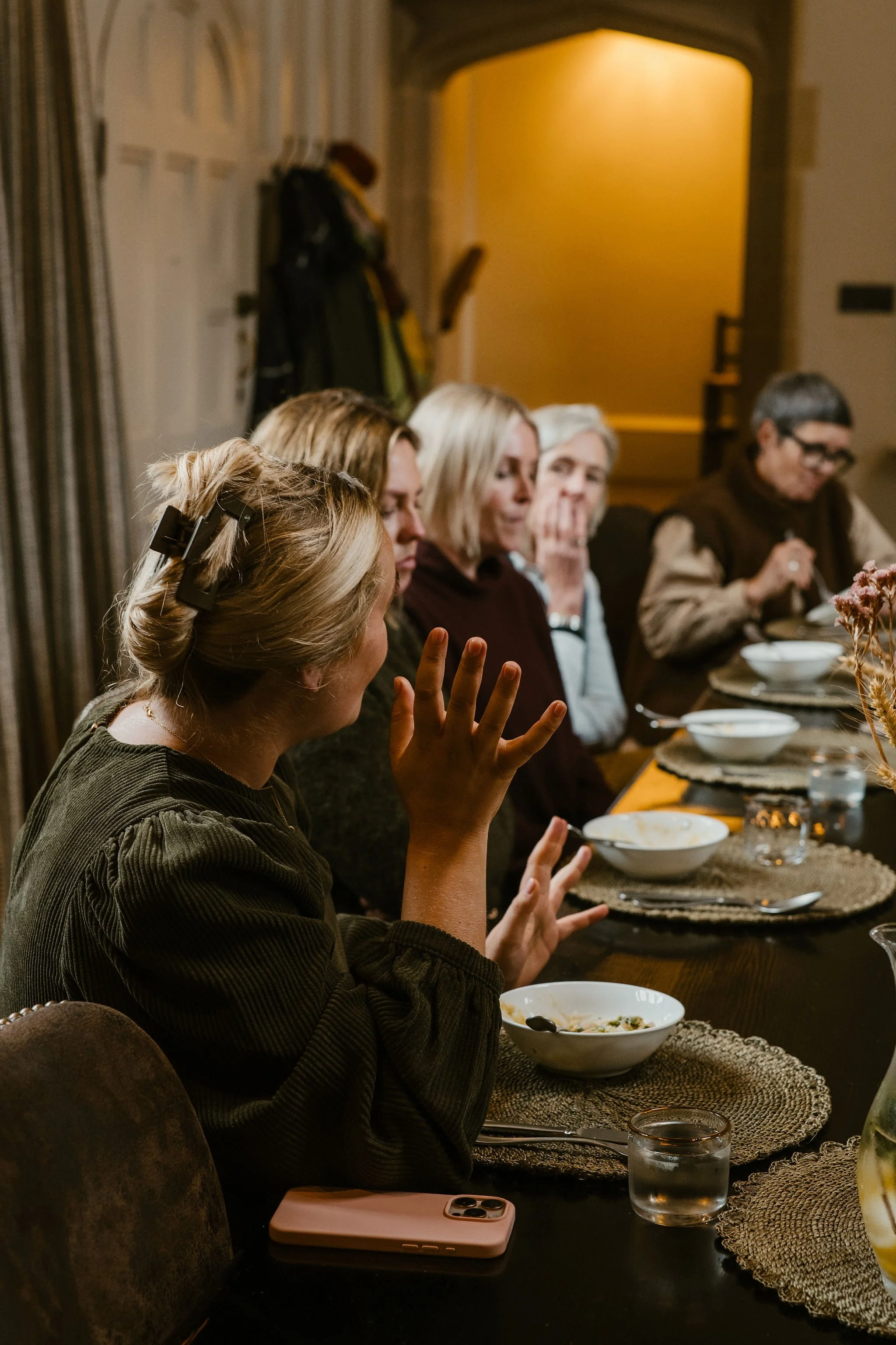 People sitting at a dining table during a meal, with one woman in the foreground gesturing with her hand.