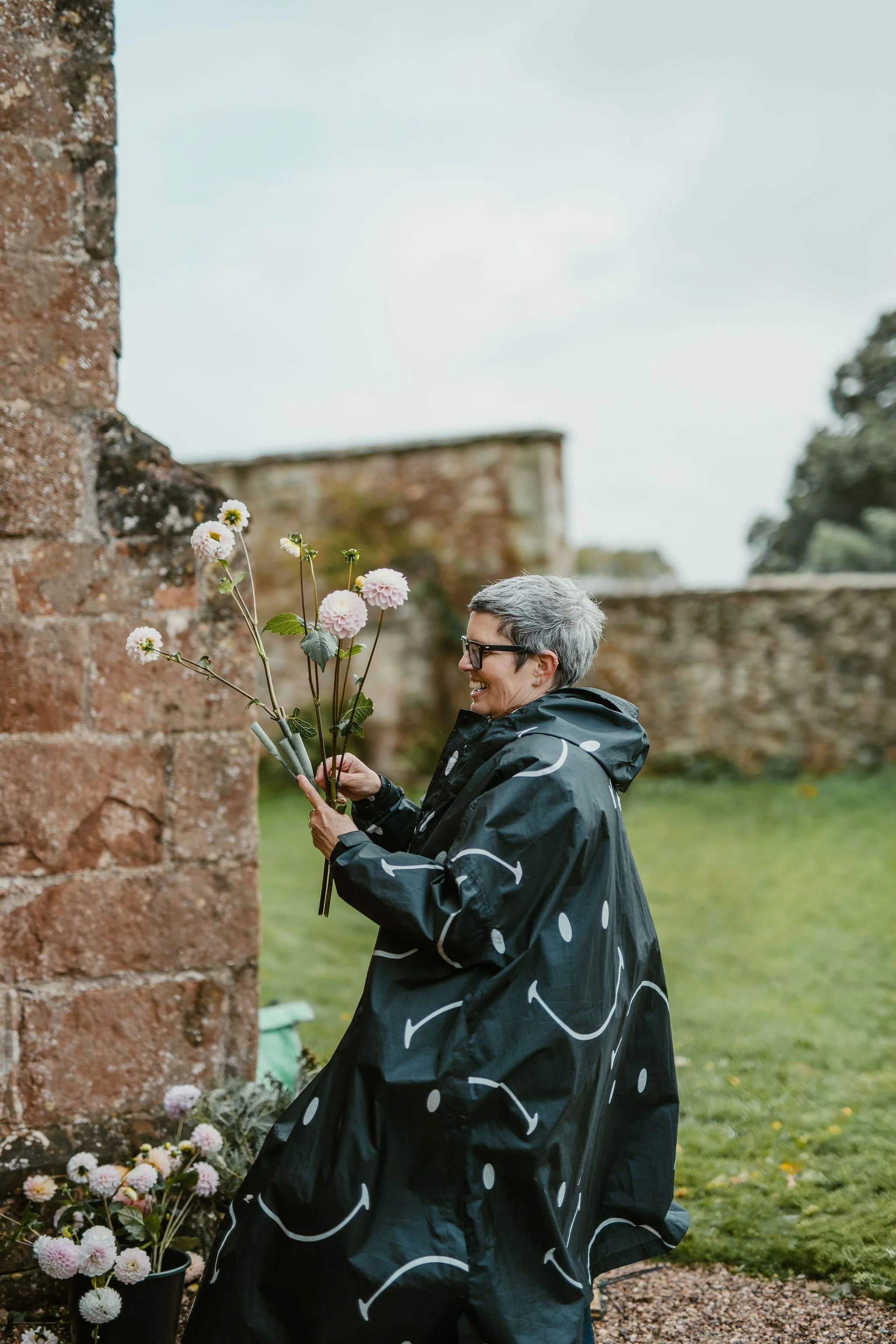 Smiling woman with short gray hair and glasses planting pink and white dahlias outside in a garden near an old brick wall on a cloudy day.