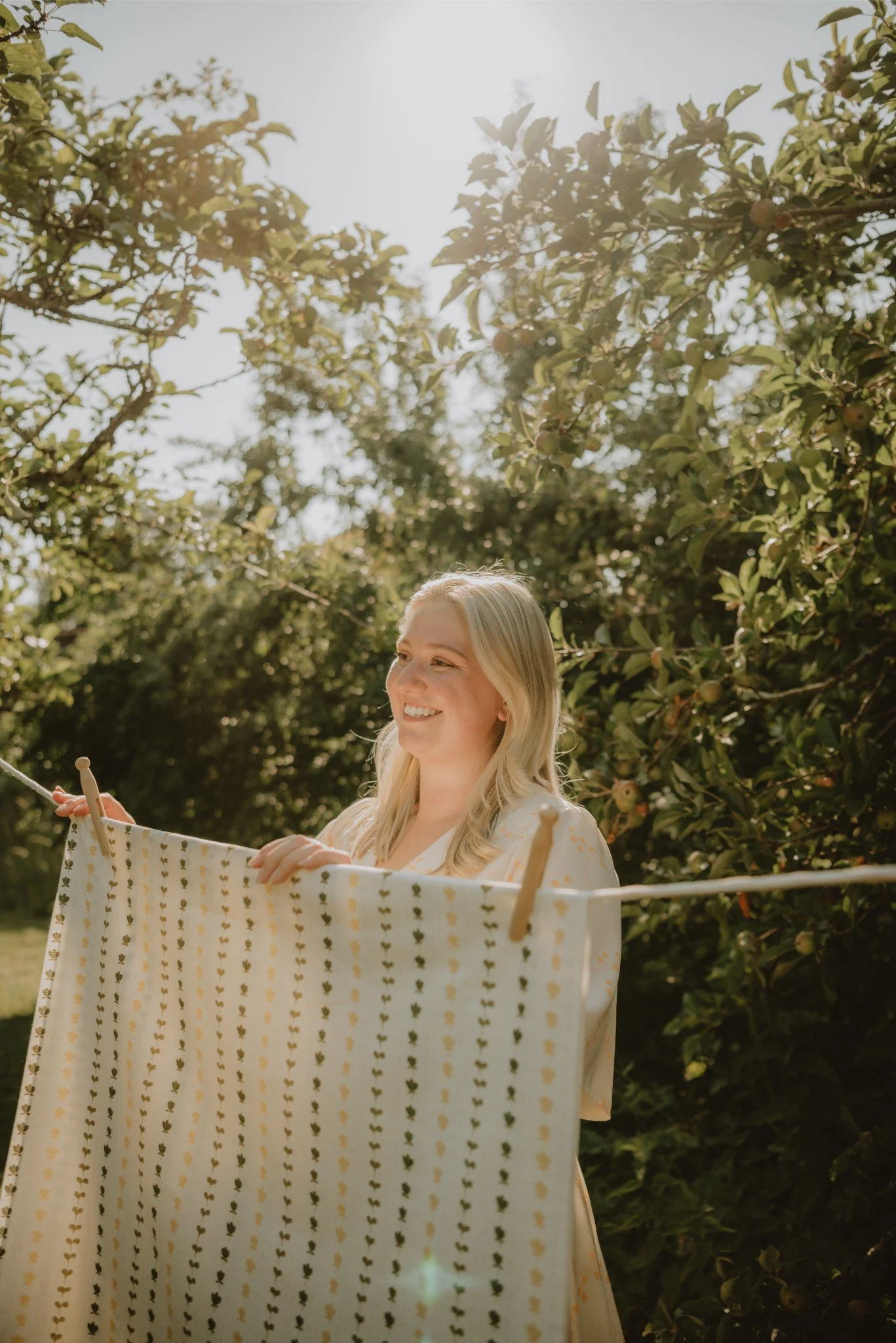 A woman with blonde hair hanging laundry on a clothesline outdoors with trees and sunlight in the background.