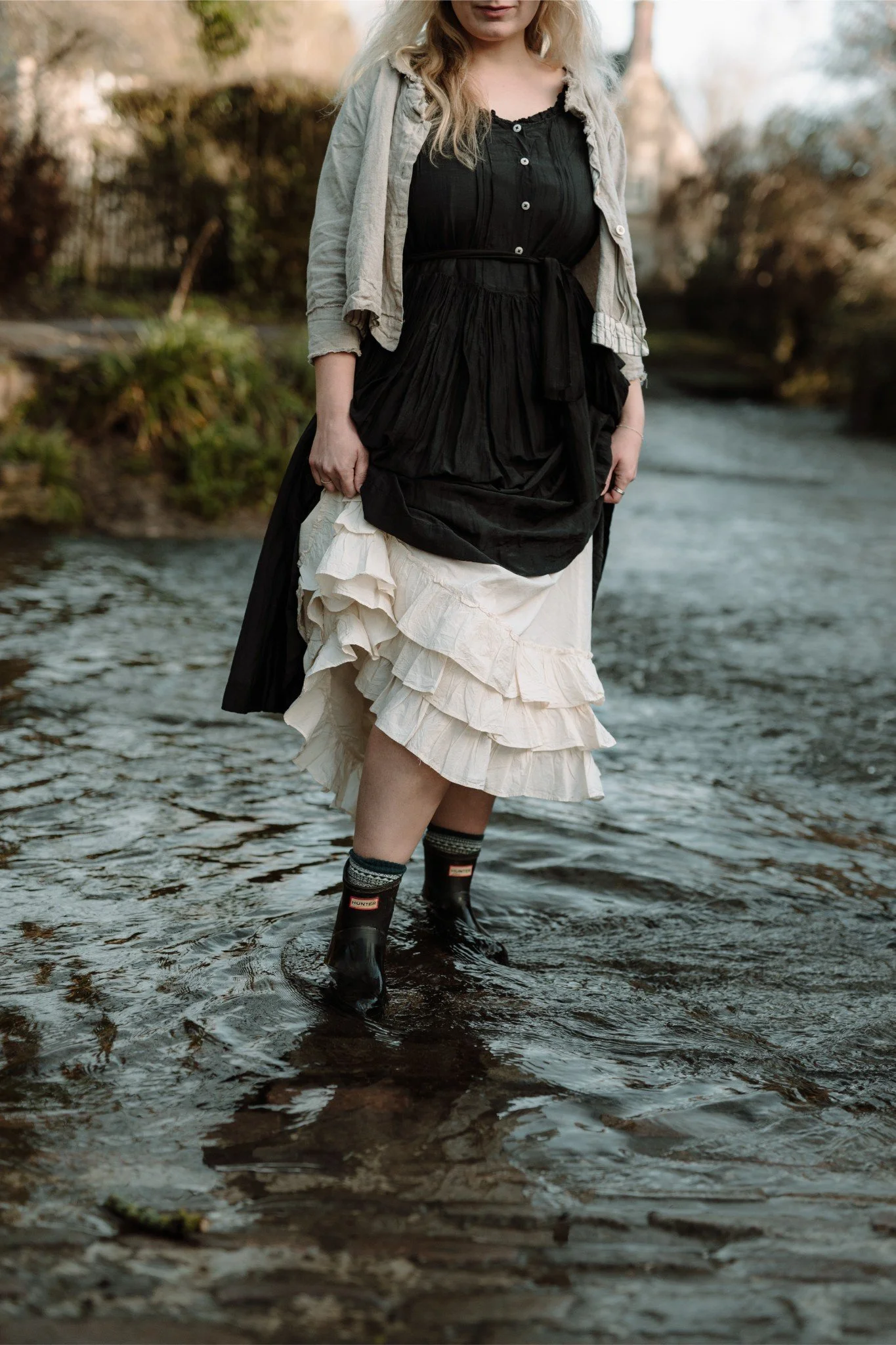 A woman standing in a stream wearing a layered skirt, black top, gray jacket, and rain boots.