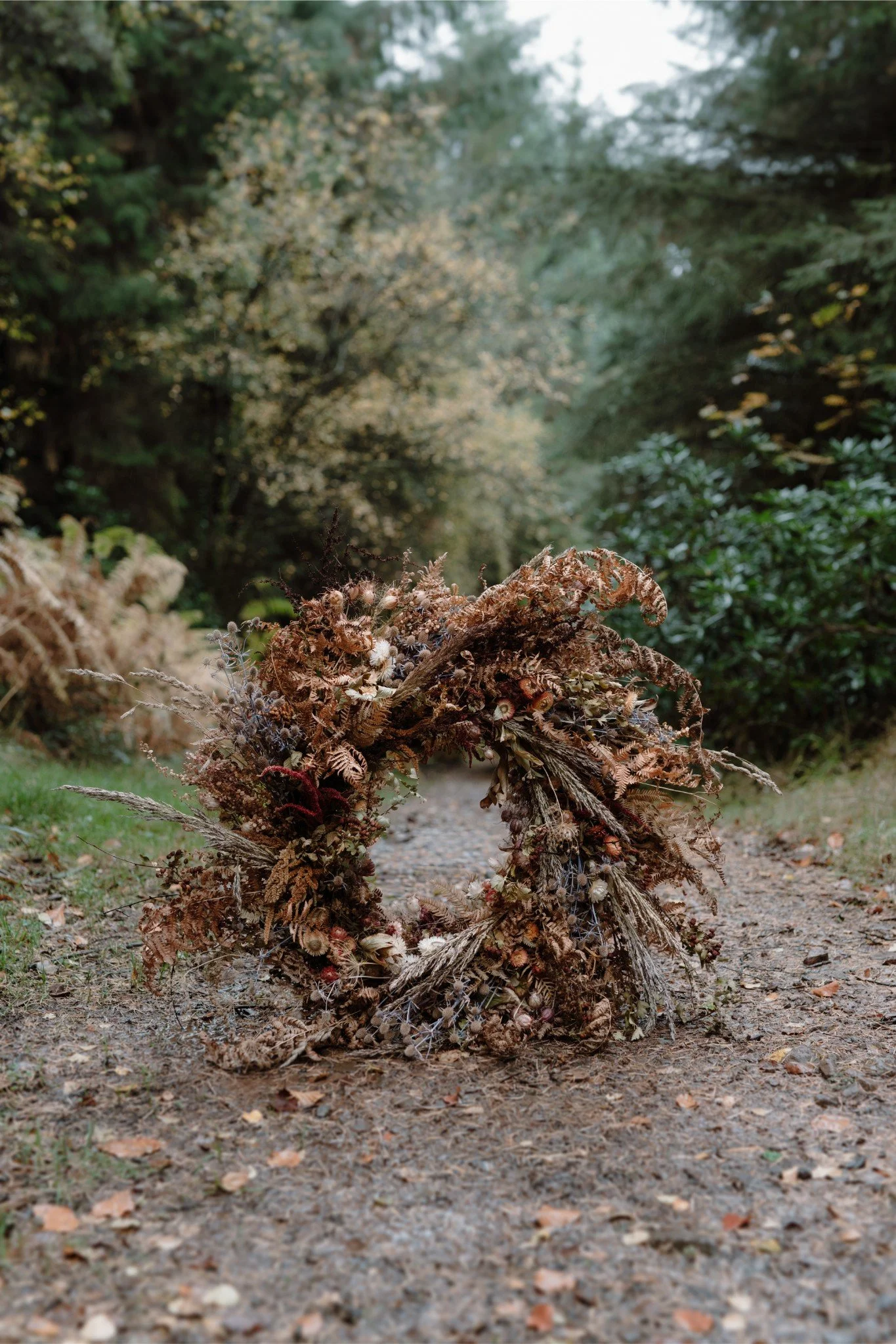 A dried flower wreath placed on a forest trail surrounded by trees with green and fall-colored leaves.