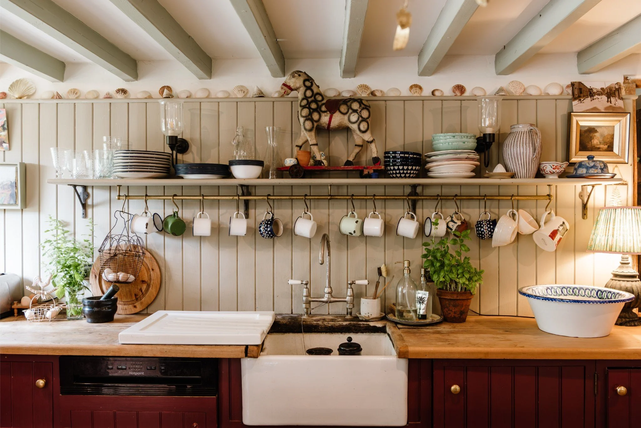 A cozy kitchen with a wooden countertop, open shelving, and a white farmhouse sink. The shelves hold plates, bowls, glasses, and decorative items. A row of cups hangs from a brass rod, and there is a potted herb plant on the counter. The walls are pa