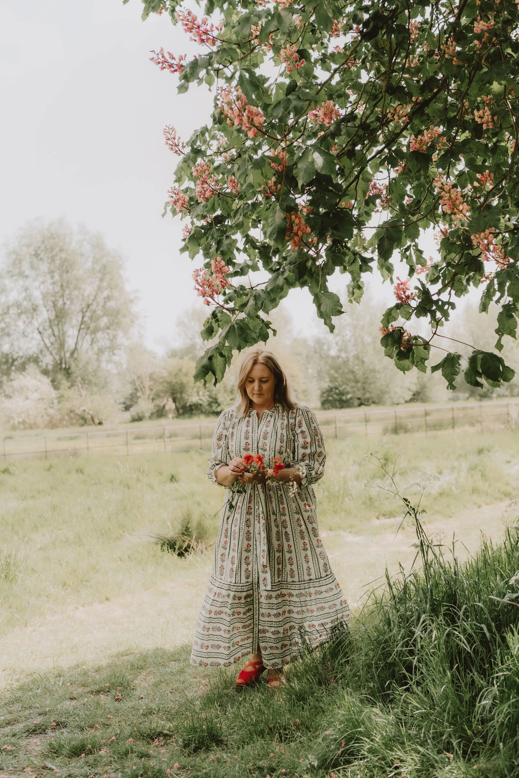 Woman standing outdoors in a grassy field, wearing a patterned dress with red shoes, holding flowers, with trees and pink blossoms overhead.