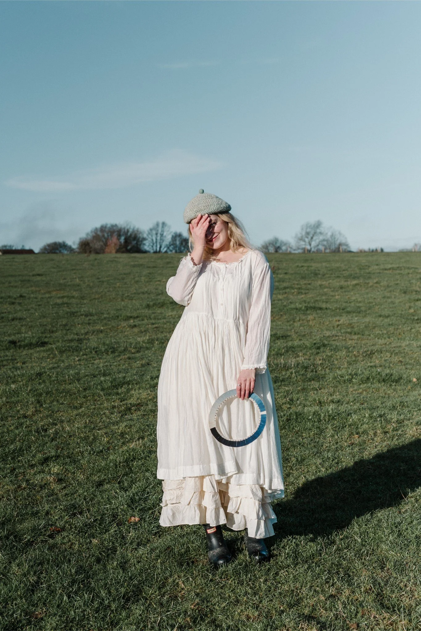 A woman in a white dress standing on grass field, wearing a grey beret, holding a circular object, covering her face with her hand, with trees and a blue sky in the background.