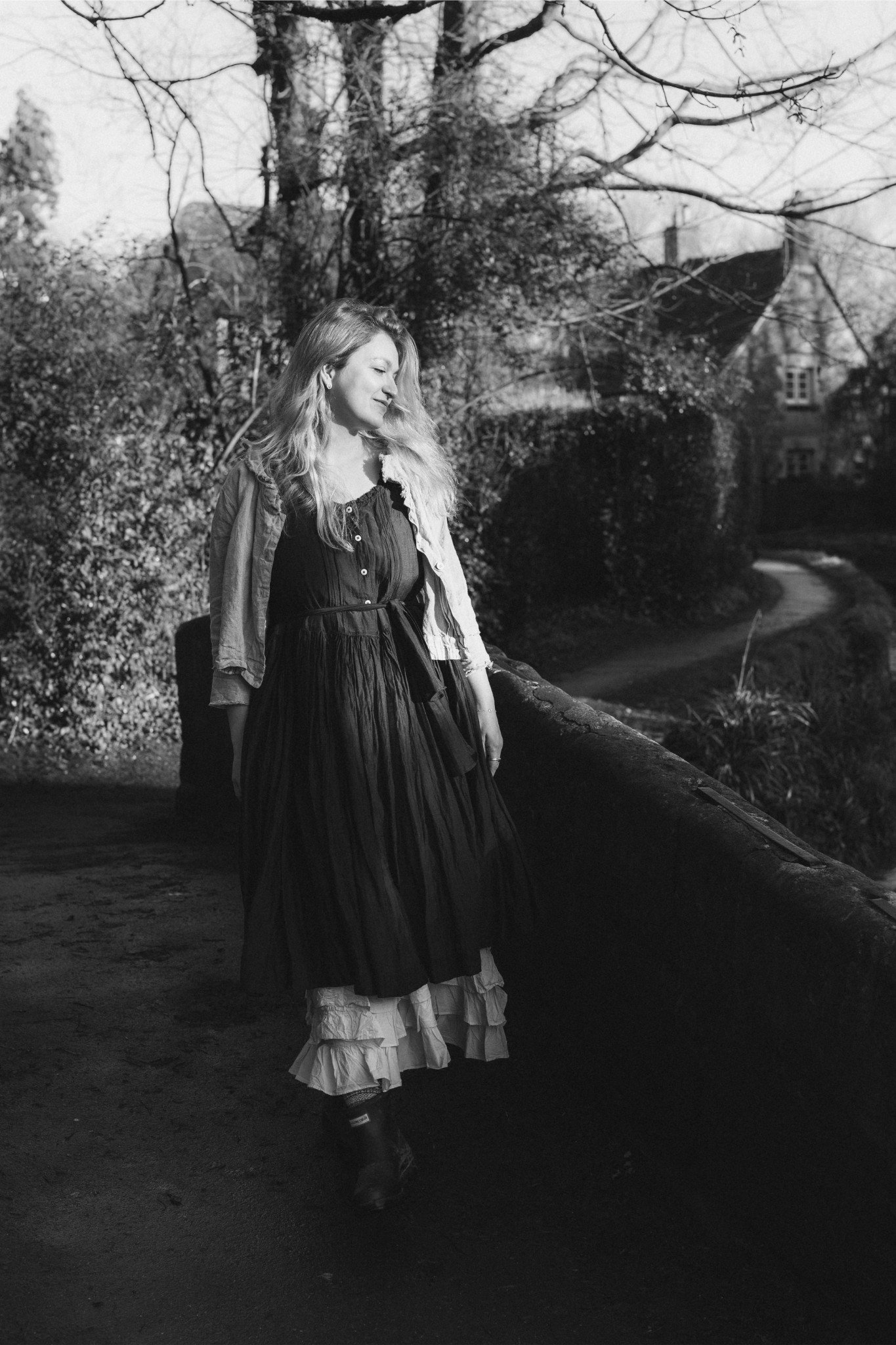 A woman with long, wavy hair wearing a layered dress and a jacket walks along a path in a park setting, with leafless trees and buildings in the background.