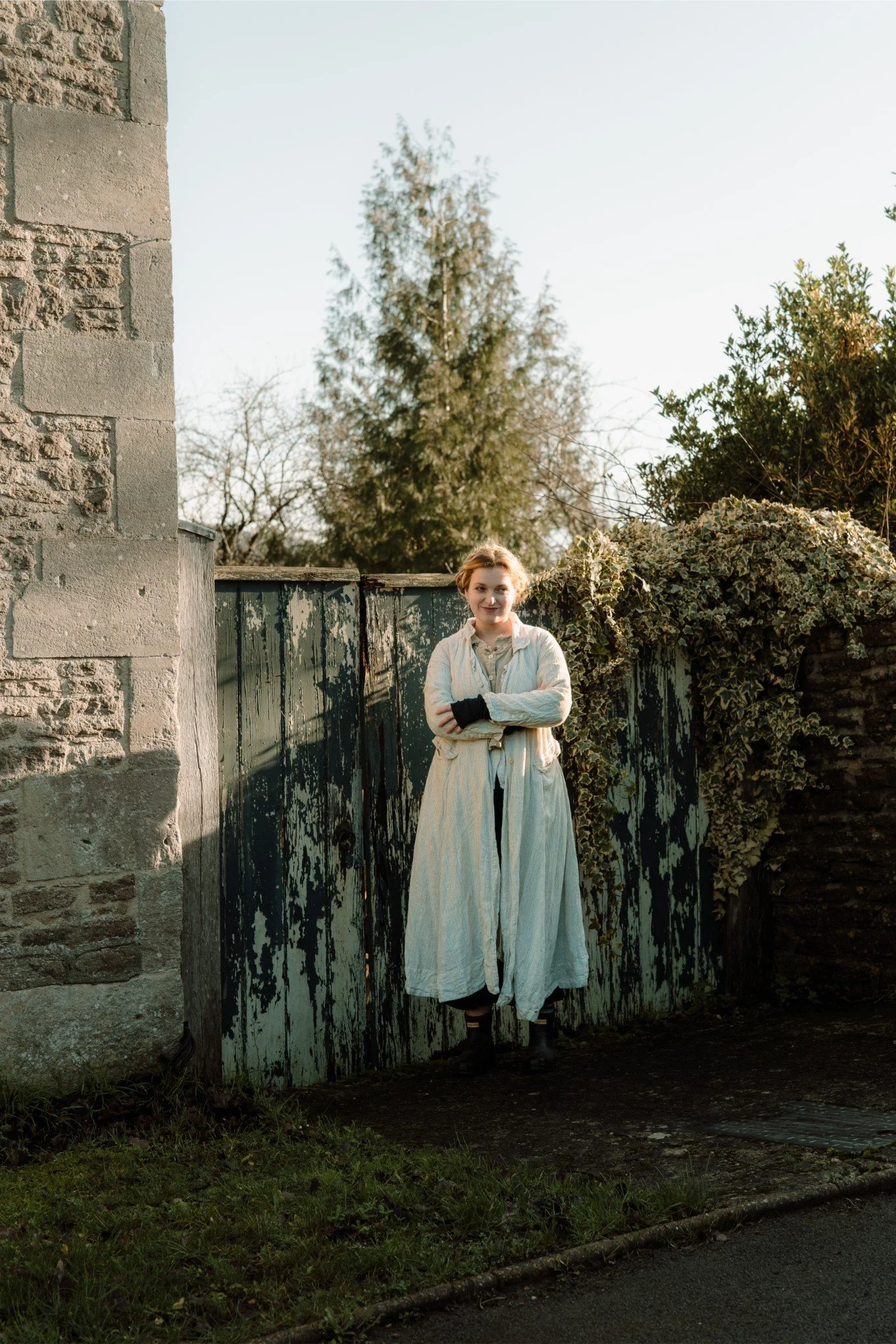 A woman with brown hair, wearing a long cream-colored dress and black boots, stands with arms crossed and smiles in front of a weathered green wooden gate, surrounded by foliage, with a large evergreen tree in the background on a clear day.