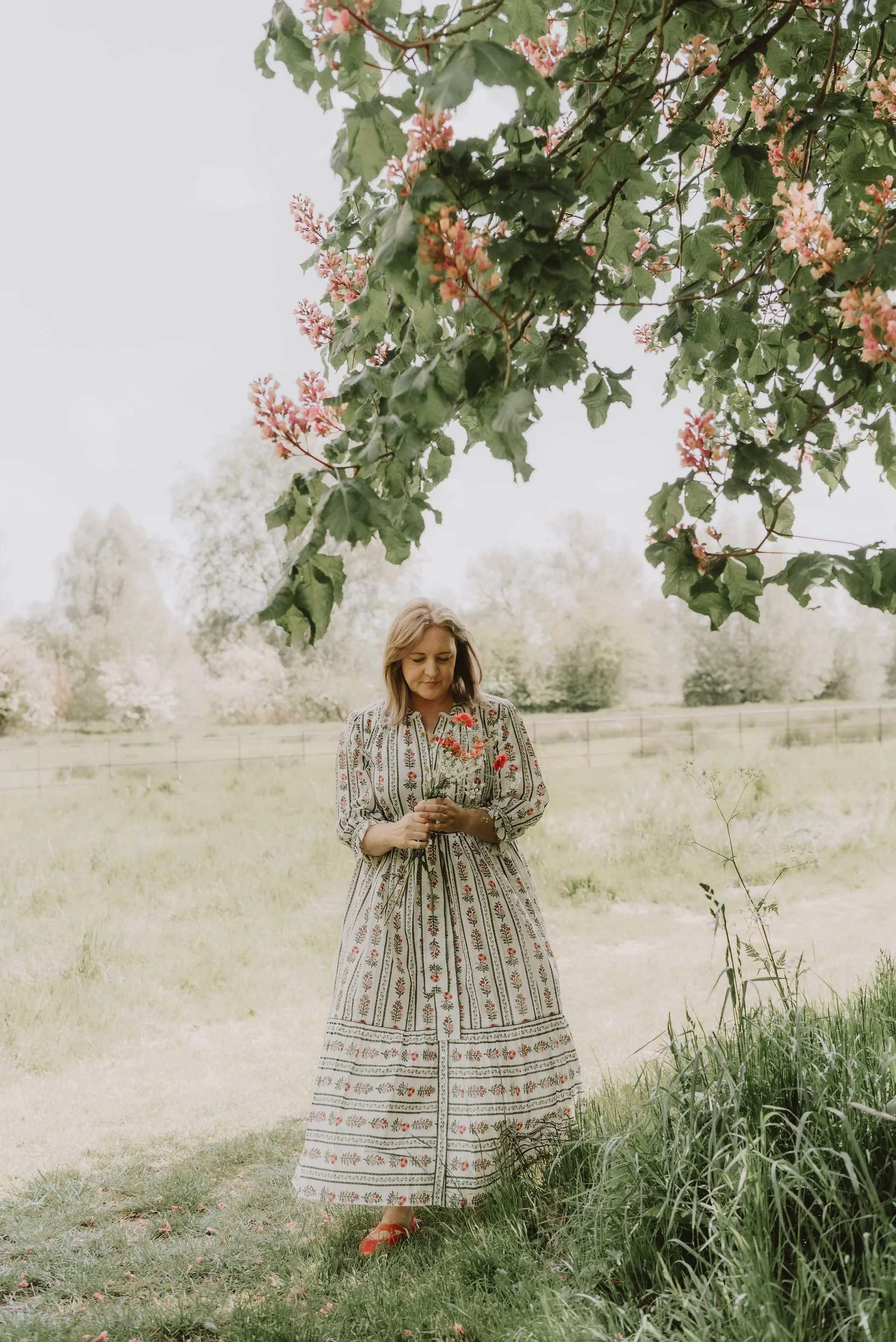 A woman is standing outdoors on a grassy field, holding a few flowers, with trees and an open sky in the background. She is wearing a long, patterned dress and red shoes, under a flowering tree with pink blossoms.