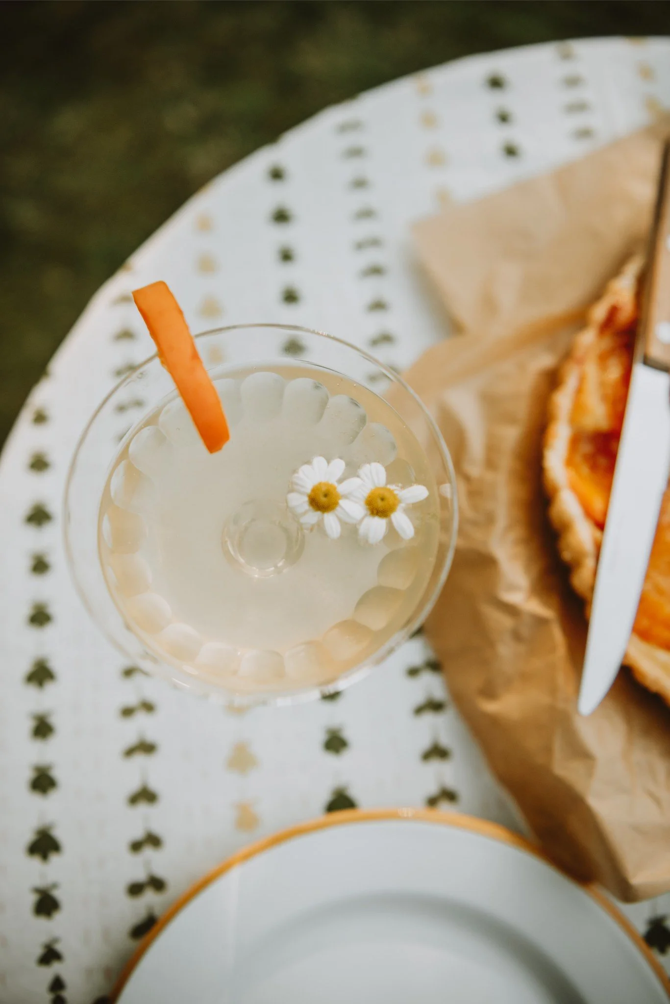 A glass of lemonade with a slice of orange and two small daisies floating on top, placed on a white table with green and gold pattern, alongside a paper-wrapped sandwich.