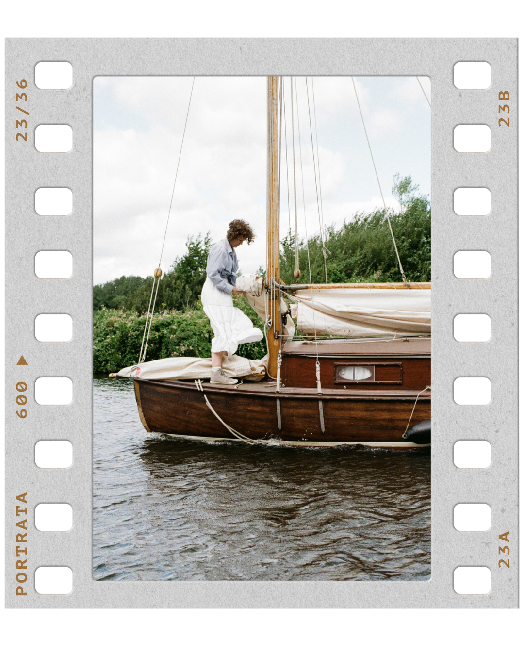 Person adjusting sails on a wooden sailboat on a river, with greenery and cloudy sky in the background.