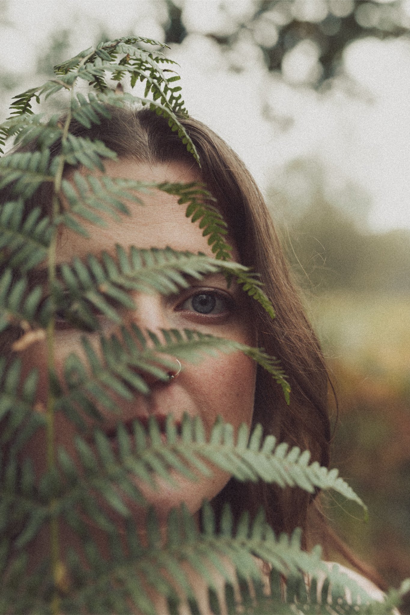 A woman with brown hair and a nose ring peeks through green fern leaves, with only one eye visible, in a natural outdoor setting.
