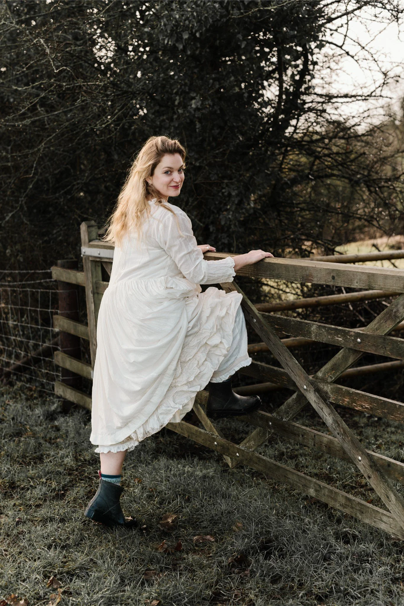A woman in a long white dress and black rain boots sitting on a wooden fence outdoors near a tree and grassy area.