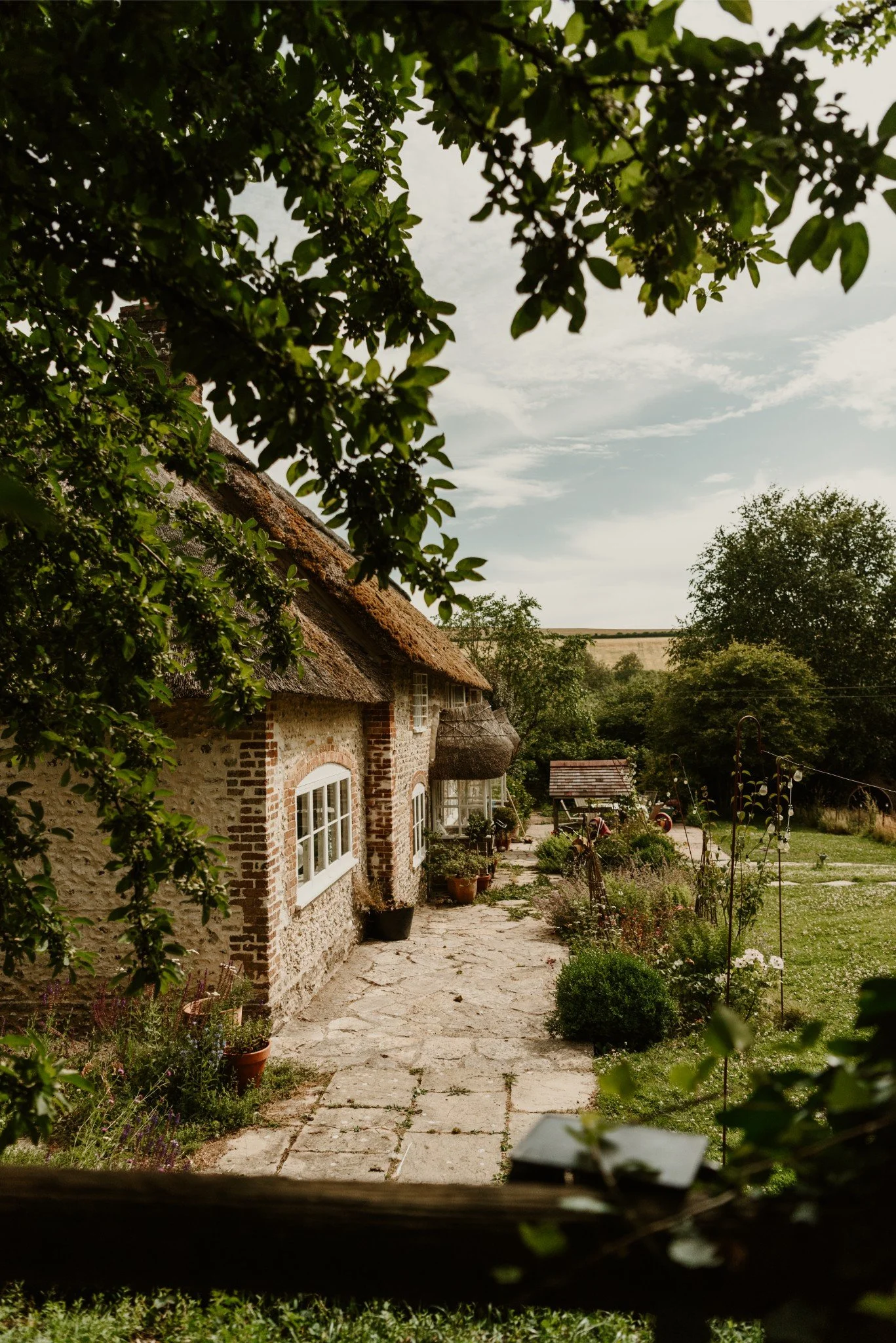 A rustic country cottage surrounded by greenery with a stone pathway leading to the entrance, trees, and a garden vine trellis in the foreground.