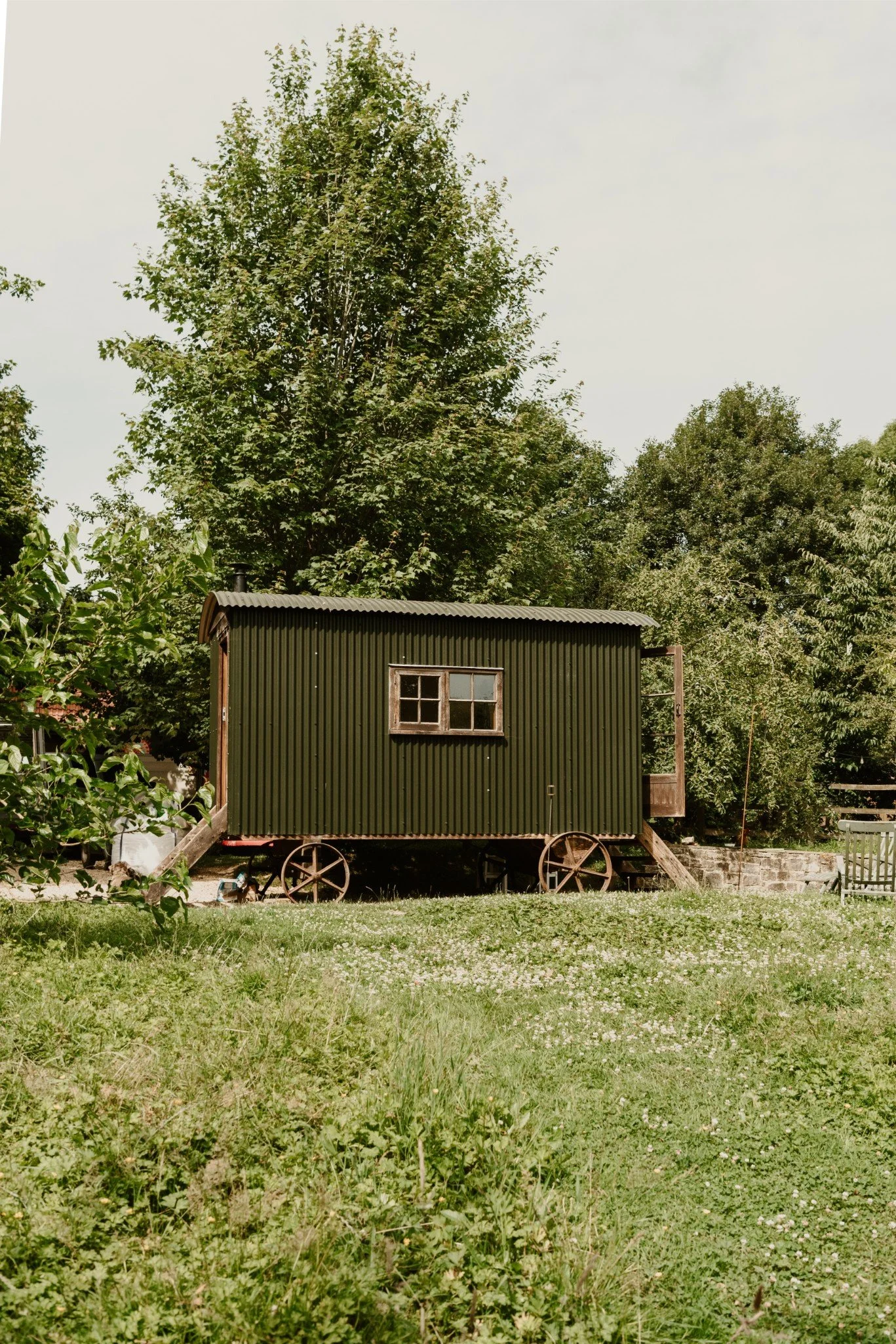 A small, dark green wooden shed on wheels with a corrugated metal roof, set in a grassy yard with trees in the background.