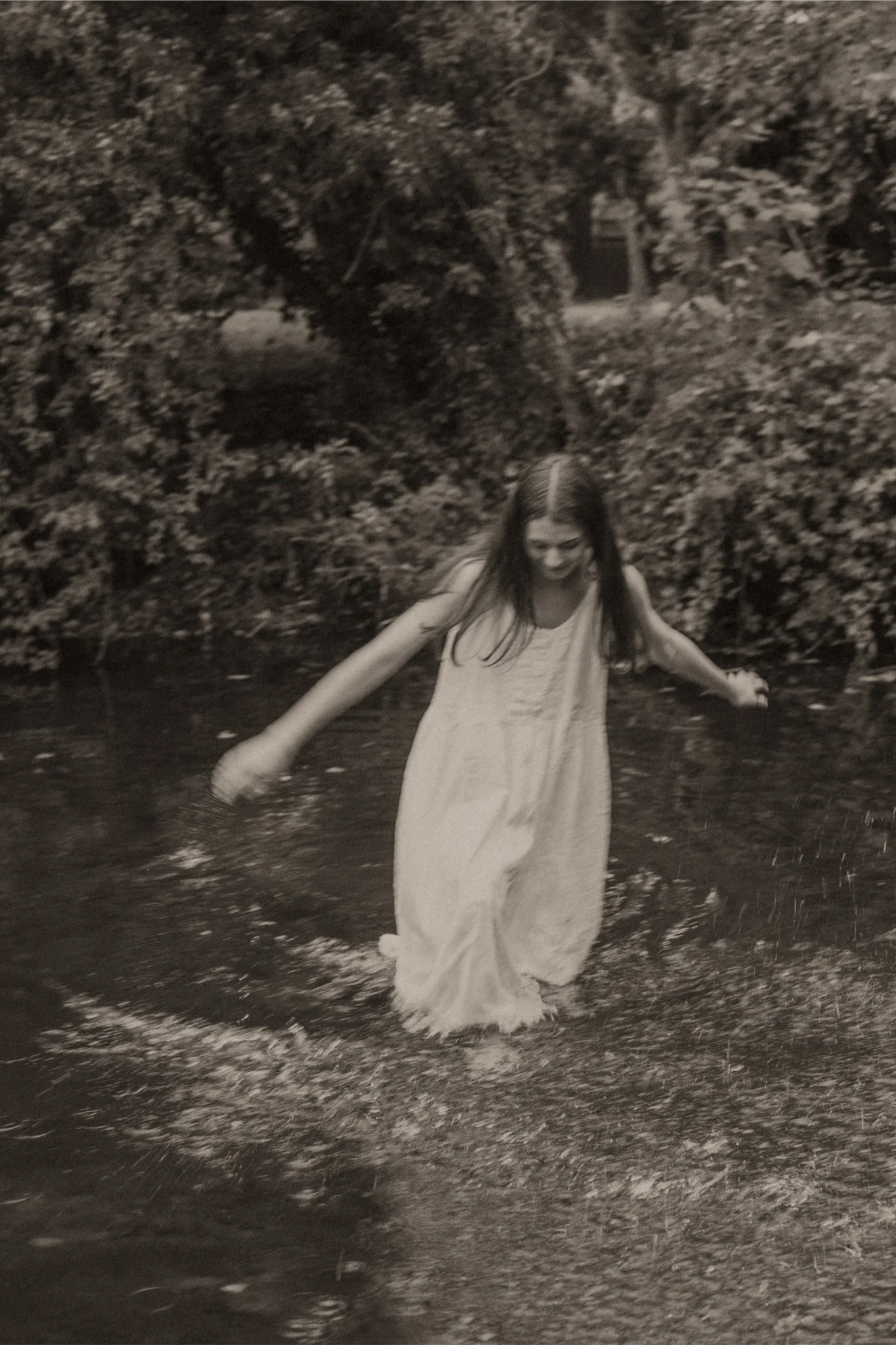 A young girl in a long dress playing and splashing in a shallow creek in a wooded area.