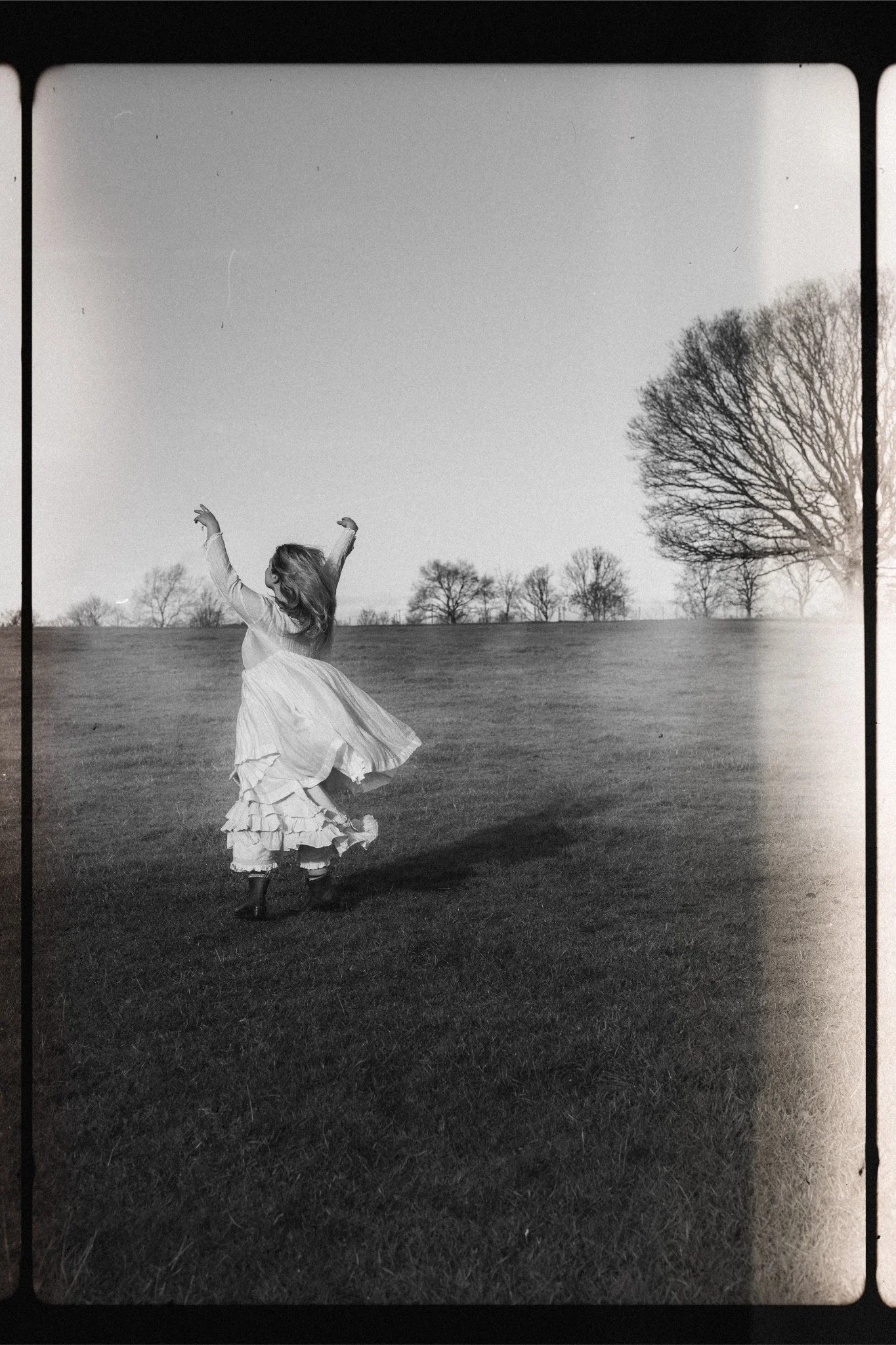A woman in a flowing dress dancing on a grassy field with trees in the background, captured in black and white.