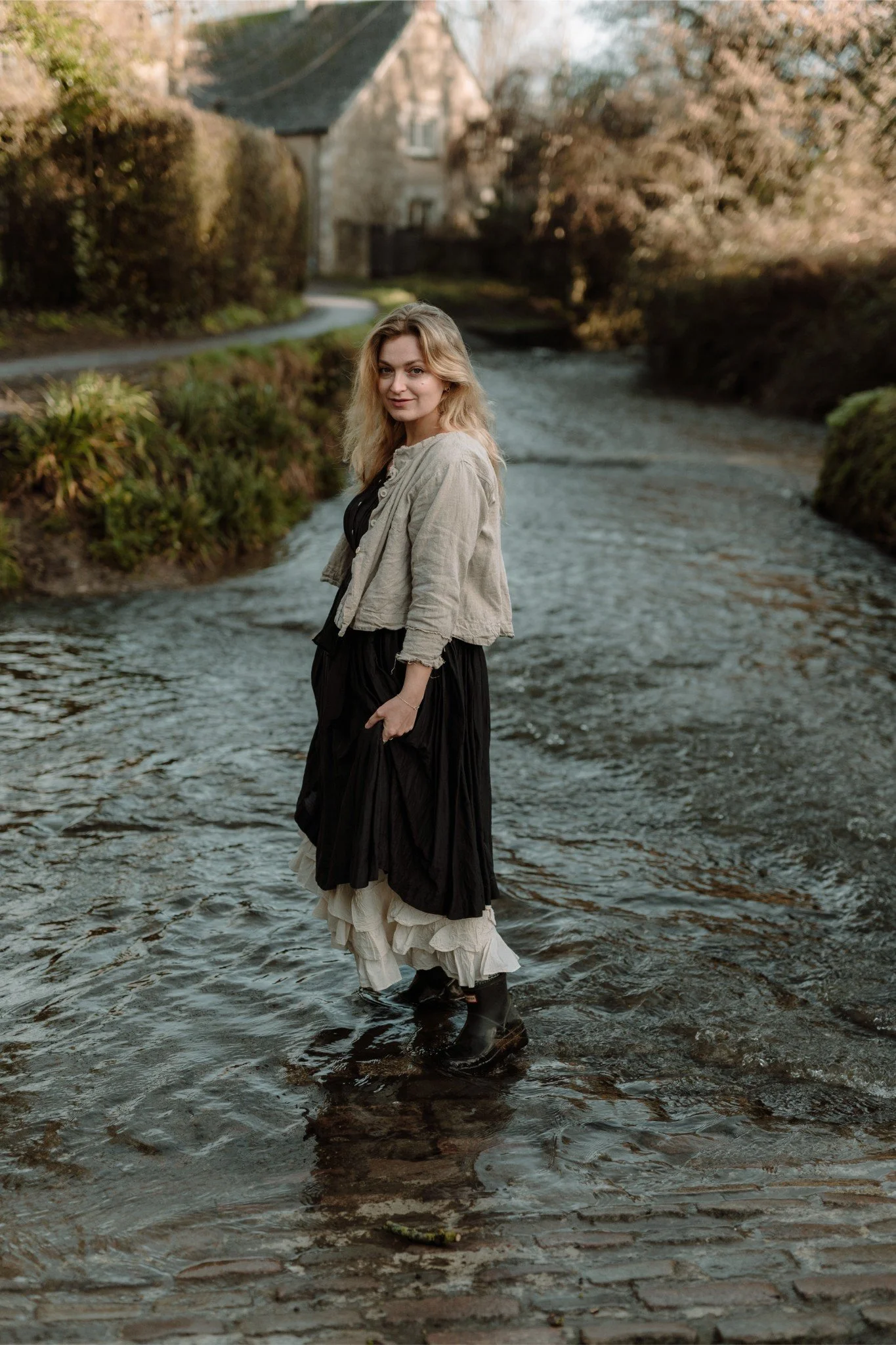 A woman standing in a shallow river with her feet in the water, surrounded by greenery and trees. She is wearing a layered dress, a beige jacket, and black boots, with a house visible in the background.