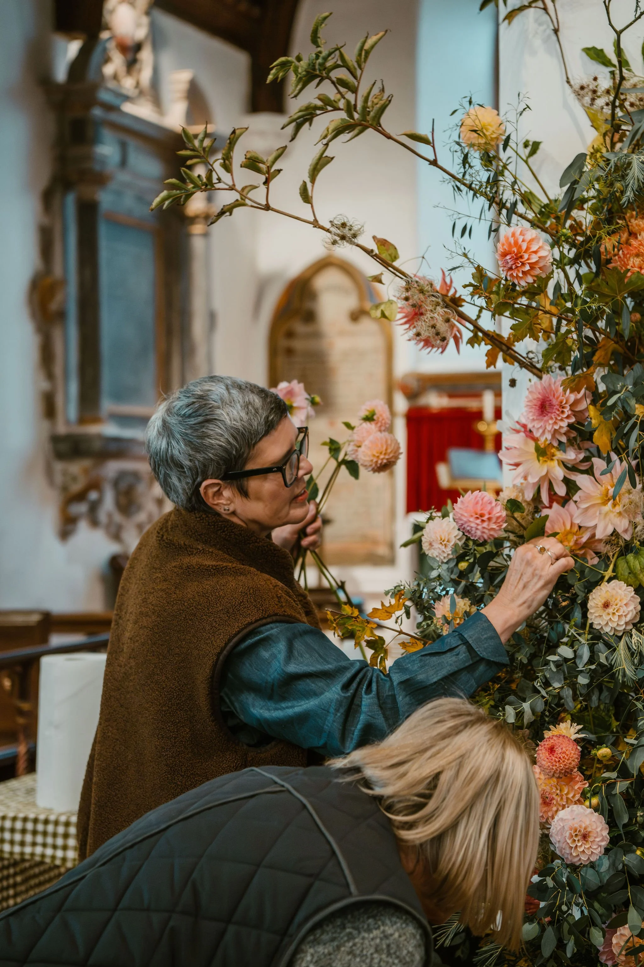 Two women arranging pink and peach flowers in a floral display inside a church or historic building.