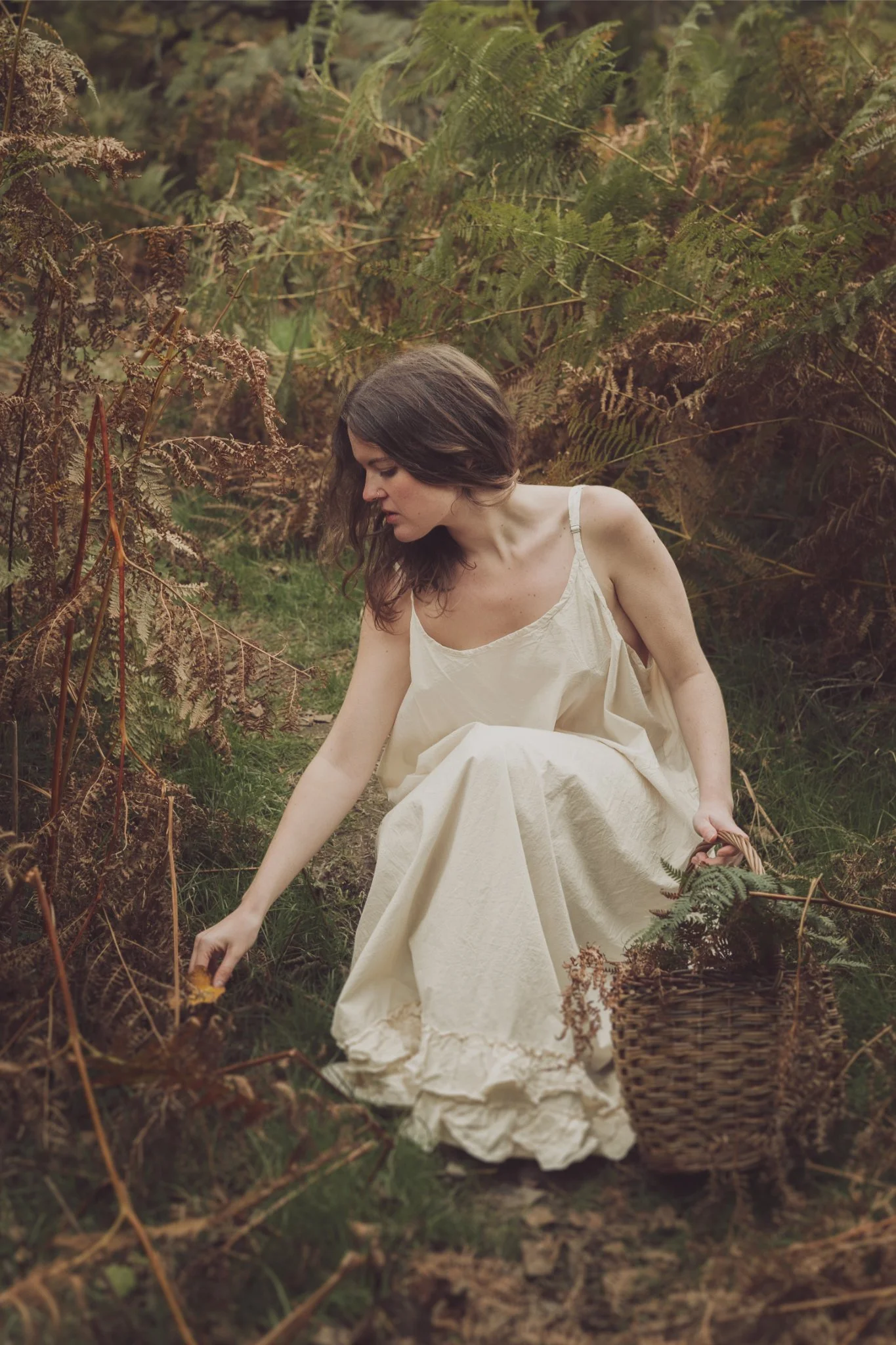 A woman in a cream-colored dress is sitting in a forest clearing surrounded by ferns, picking a yellow leaf from the ground and holding a woven basket.
