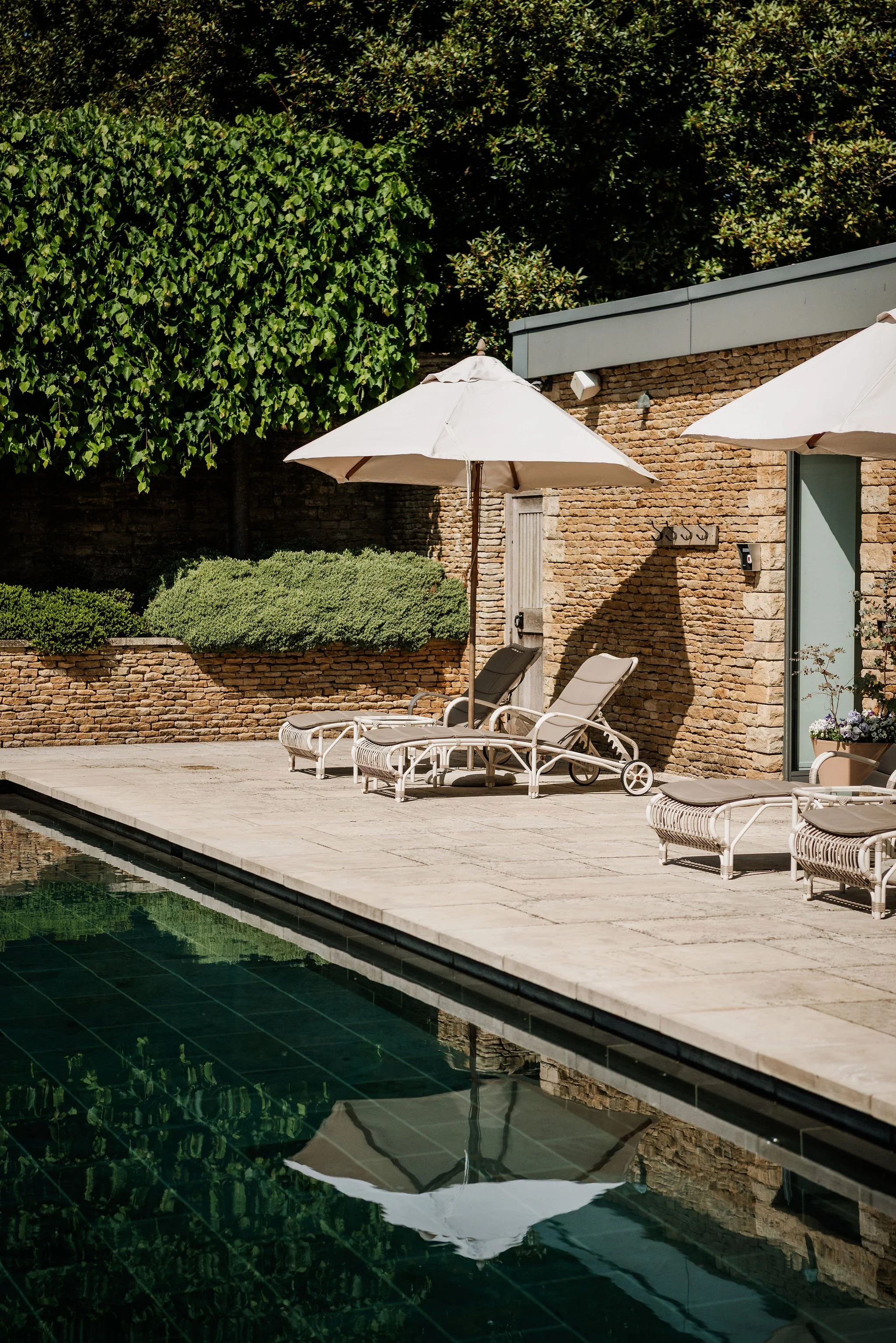 Poolside scene with two lounge chairs under white umbrellas, a brick wall, green bushes, and trees reflected in the pool water.