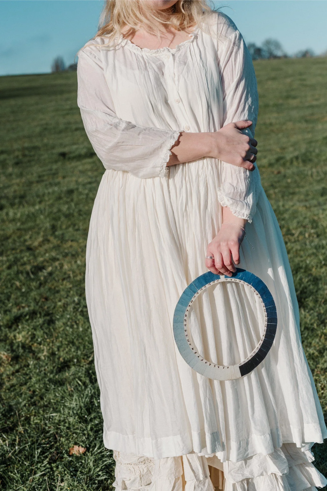 A woman in a white, pleated, vintage-style dress standing outdoors on grass, holding a circular color palette in one hand, with the sky and distant trees in the background.