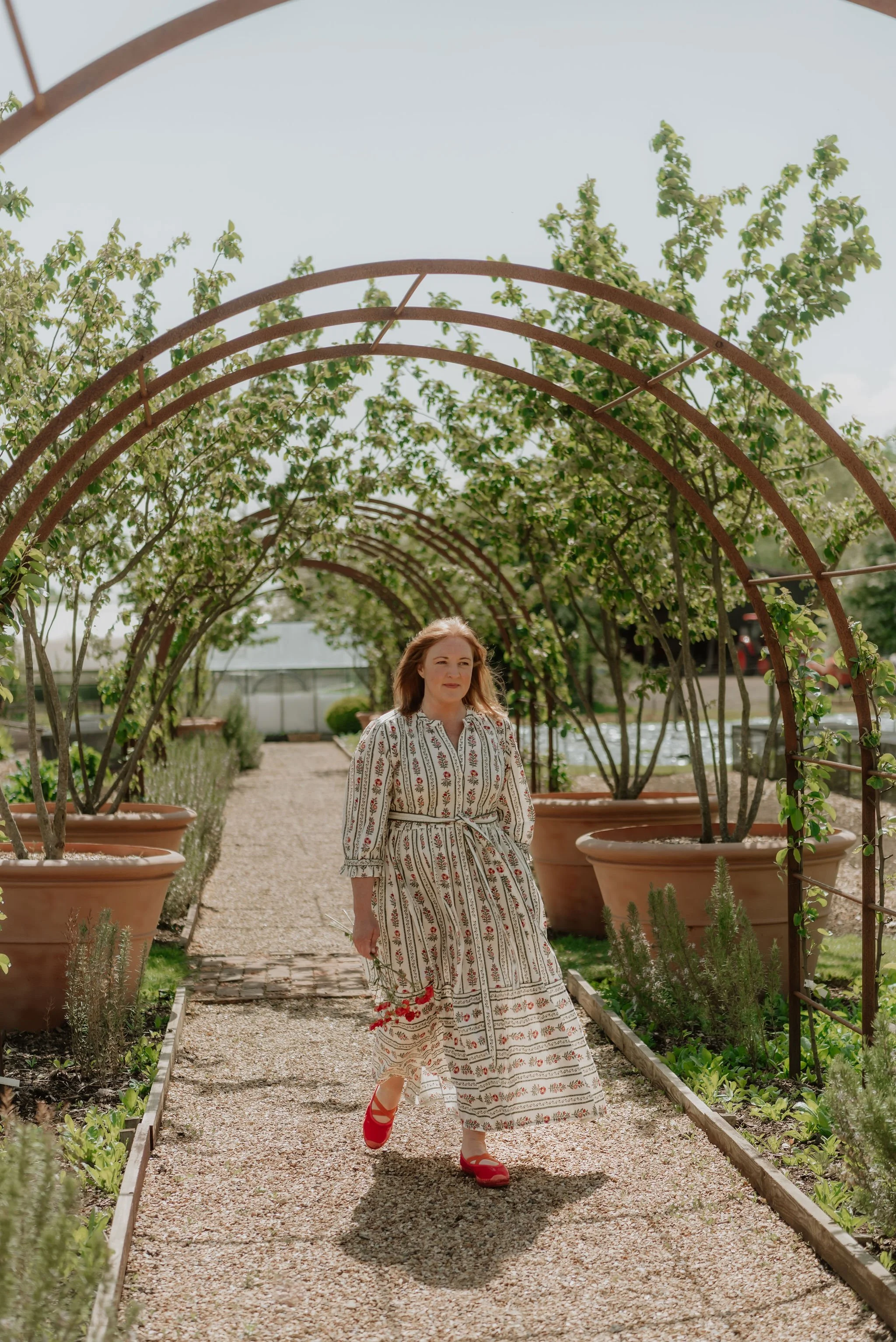 A woman walking through a garden with arched metal structures covered in green plants and flowers, wearing a patterned long dress and red shoes.