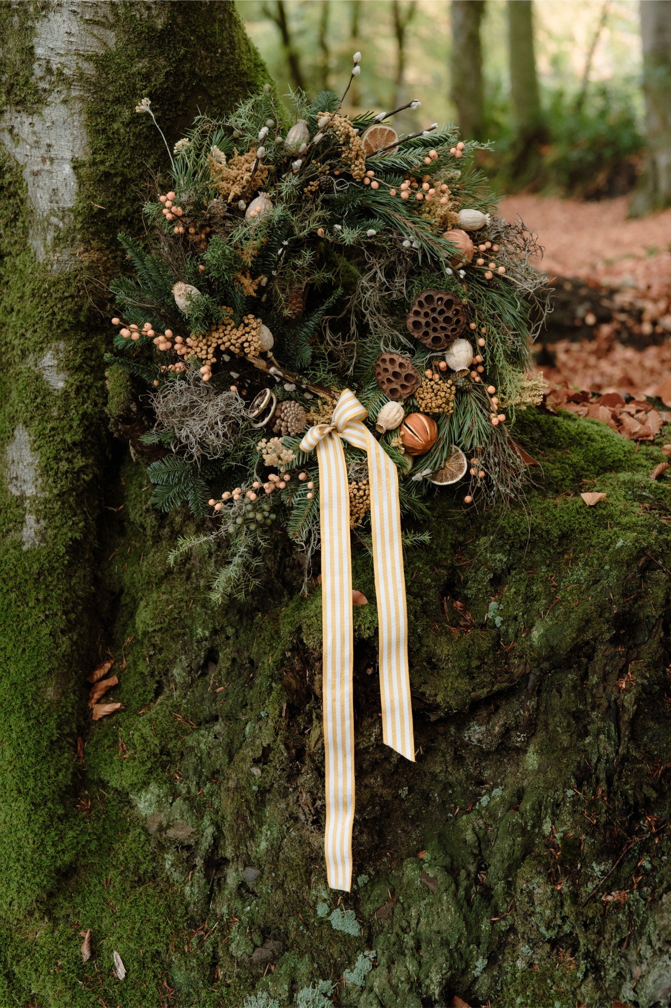 A decorative wreath made of greenery, dried flowers, seed pods, and natural elements, hanging on a moss-covered tree trunk in a forest setting.