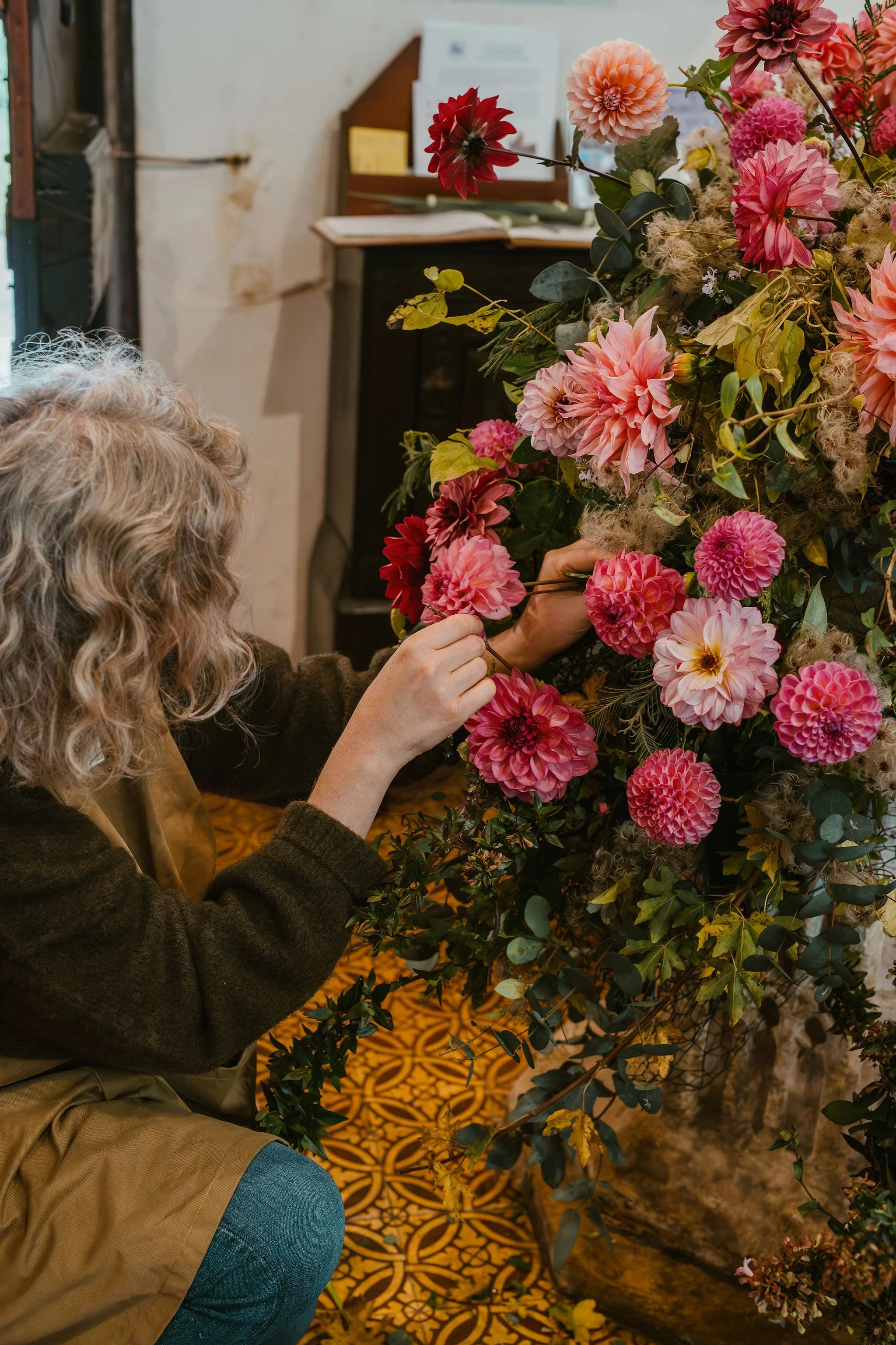 A woman with curly hair adjusting pink and red dahlias and other flowers in a floral arrangement.