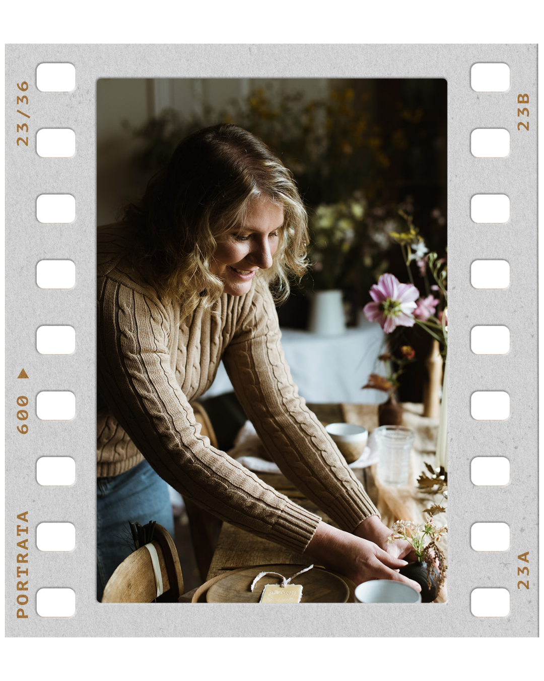 A woman in a beige sweater arranging flowers on a table in a cozy, decorated room.