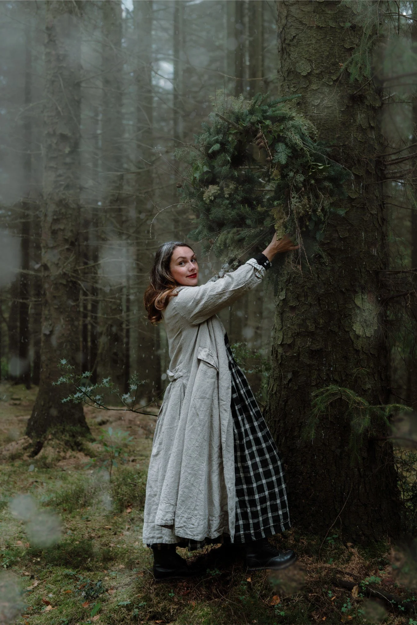 A woman dressed in vintage clothing, including a checkered skirt and beige coat, stands in a forest holding a Christmas wreath in front of a large tree.