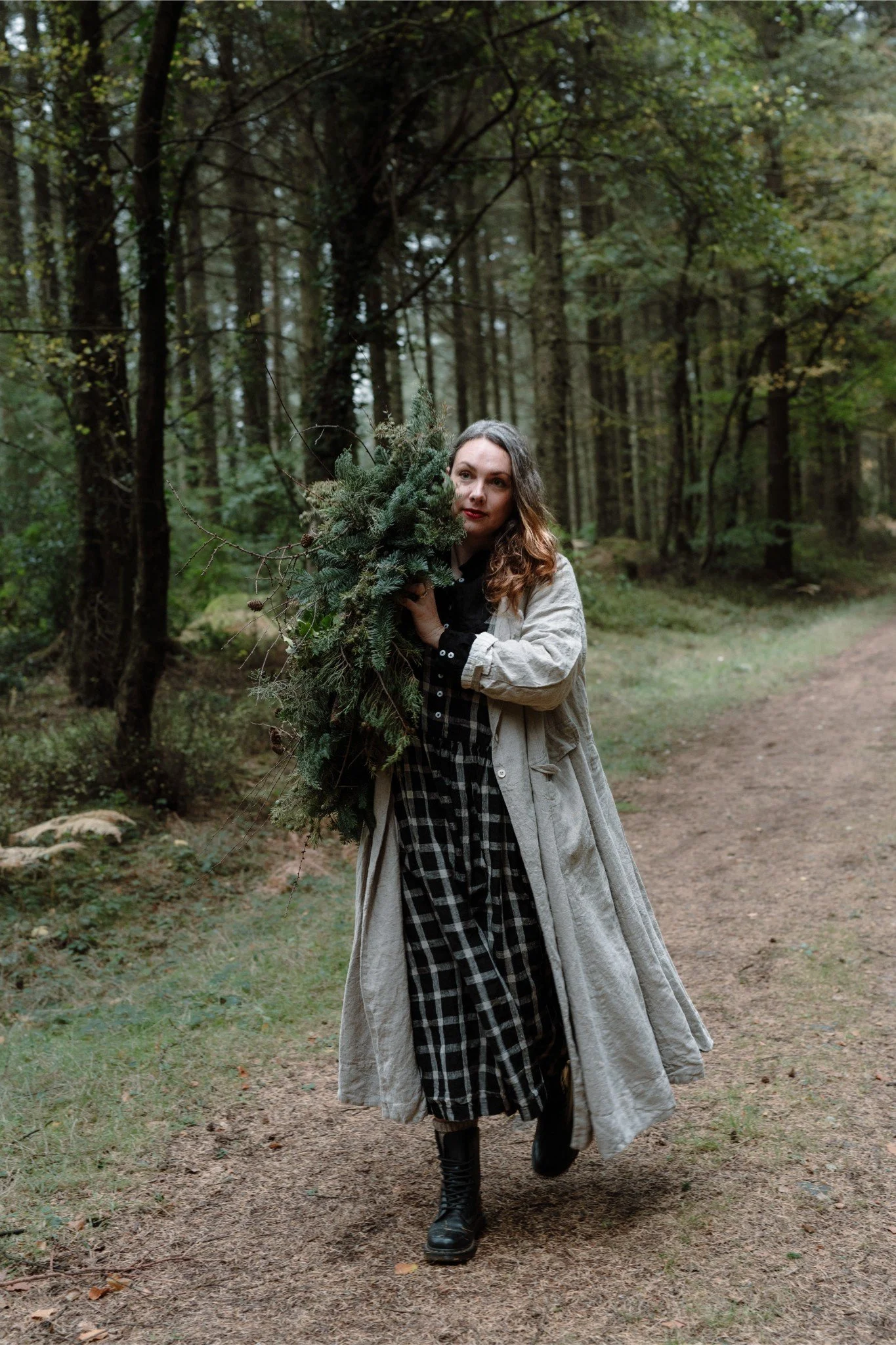 A woman walking in a forest holding a large bundle of pine branches and greenery.