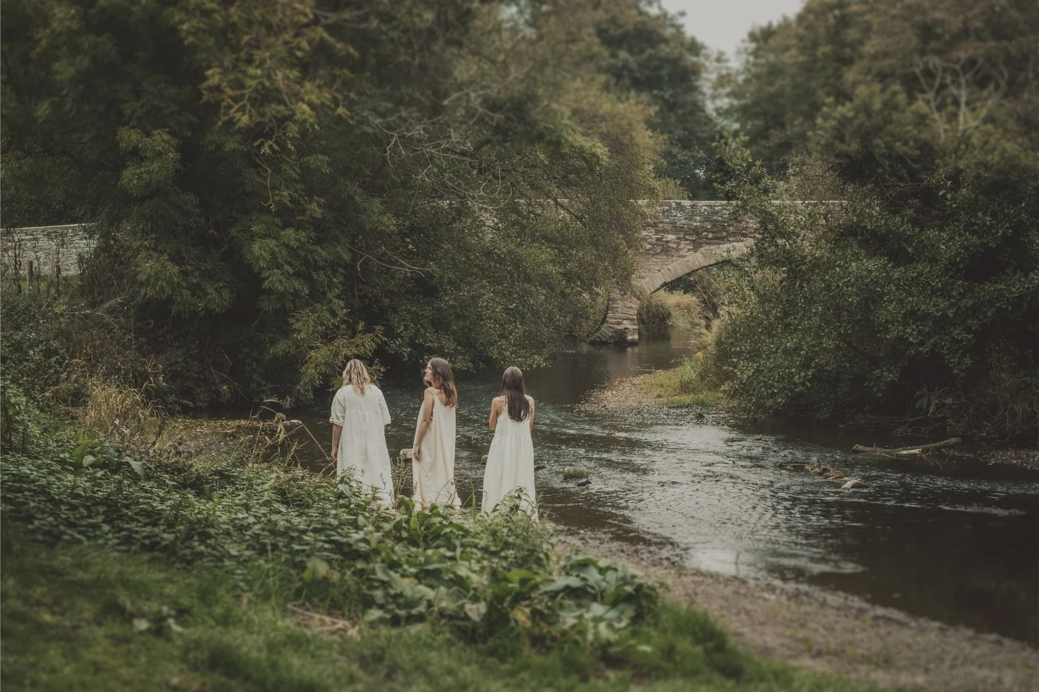 Three women in white dresses standing by a river with lush green trees and an old stone bridge in the background during daytime.
