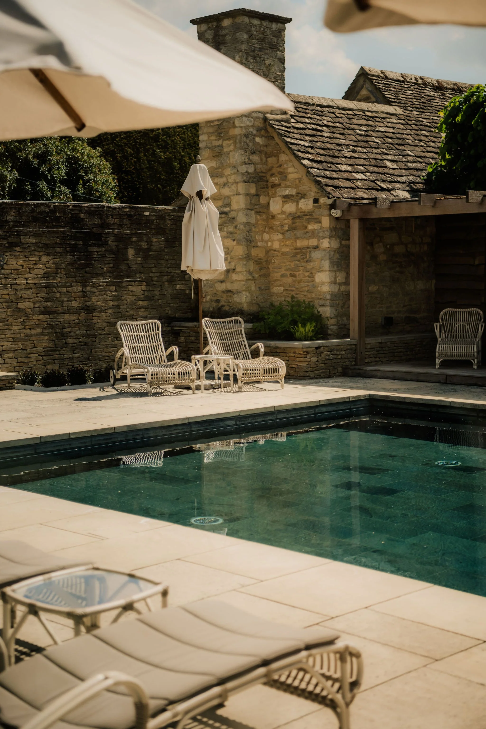 A private outdoor swimming pool surrounded by beige stone tiles, with white lounge chairs and umbrellas. In the background, there is a stone building with a sloped tiled roof and a brick chimney, and a tall stone wall covered with greenery.