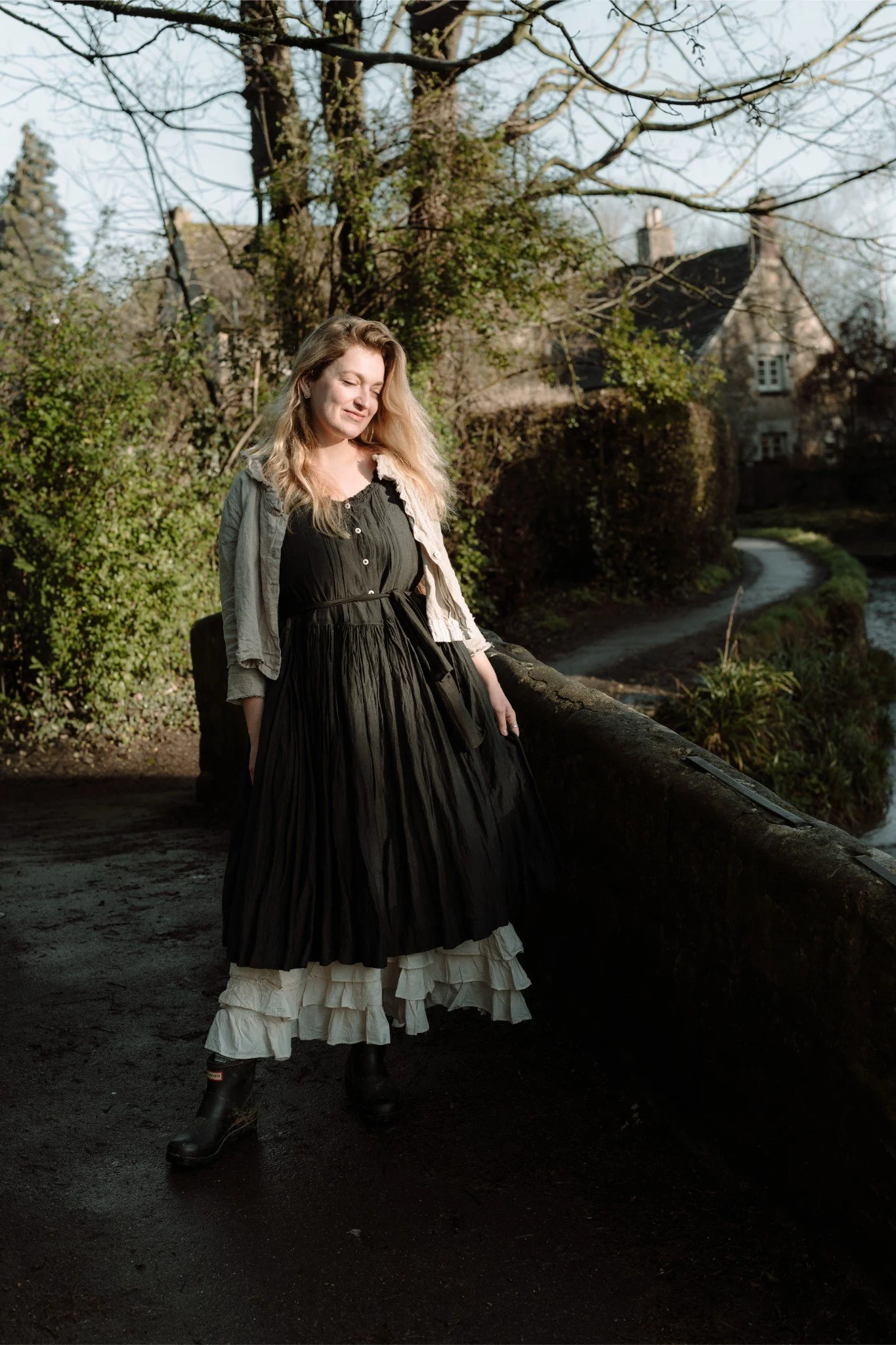A woman standing outdoors on a paved path, wearing a black dress and cream-colored ruffled petticoat, with a light jacket and boots, next to a stone wall with bushes and trees in the background.
