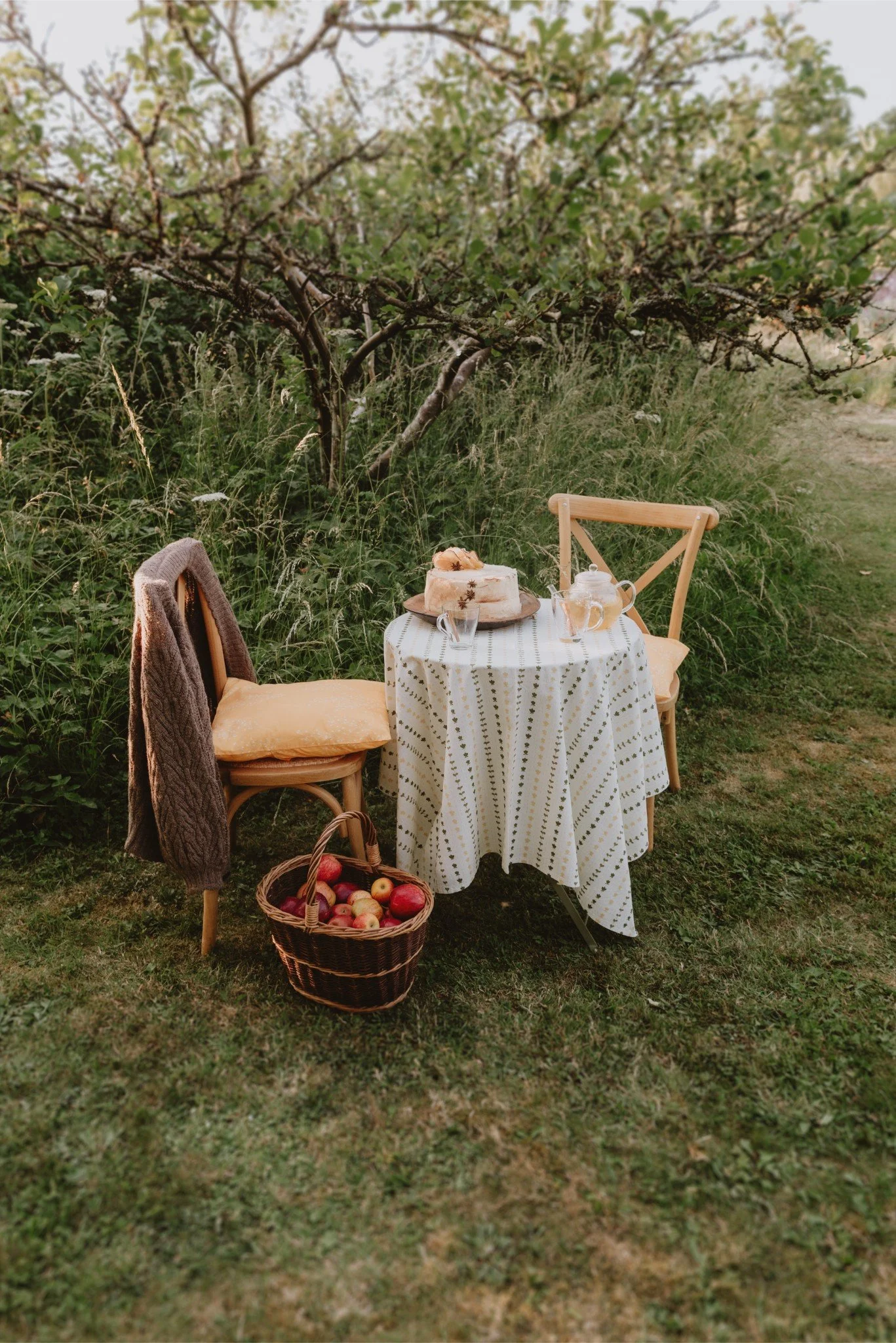 An outdoor breakfast scene with a small round table covered with a white tablecloth with gold dot patterns. On the table is a cake, a glass teapot, and a glass. Two wooden chairs are positioned around the table, one has a brown fur coat draped over t
