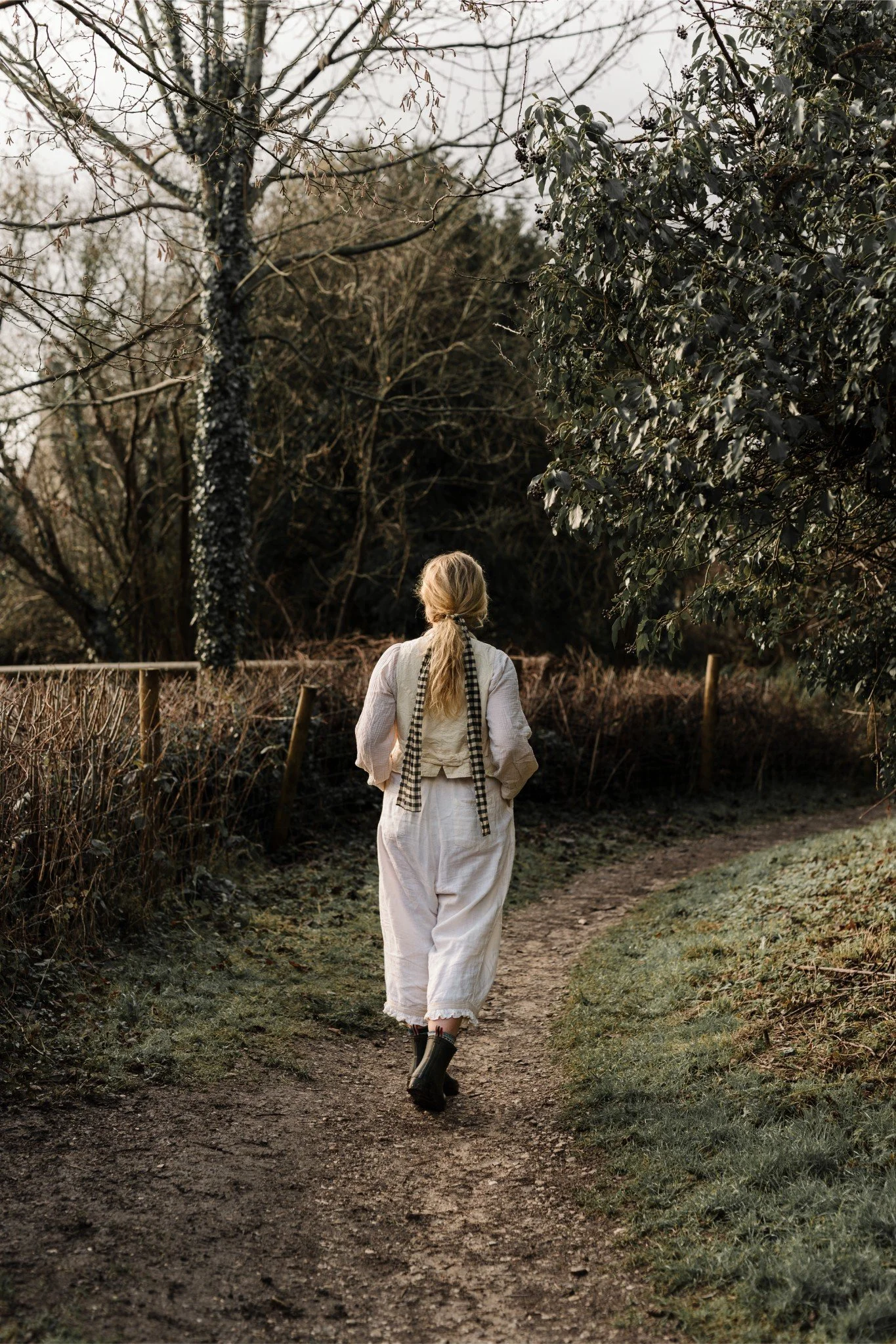 A person with long blonde hair walking on a dirt path through a wooded area.