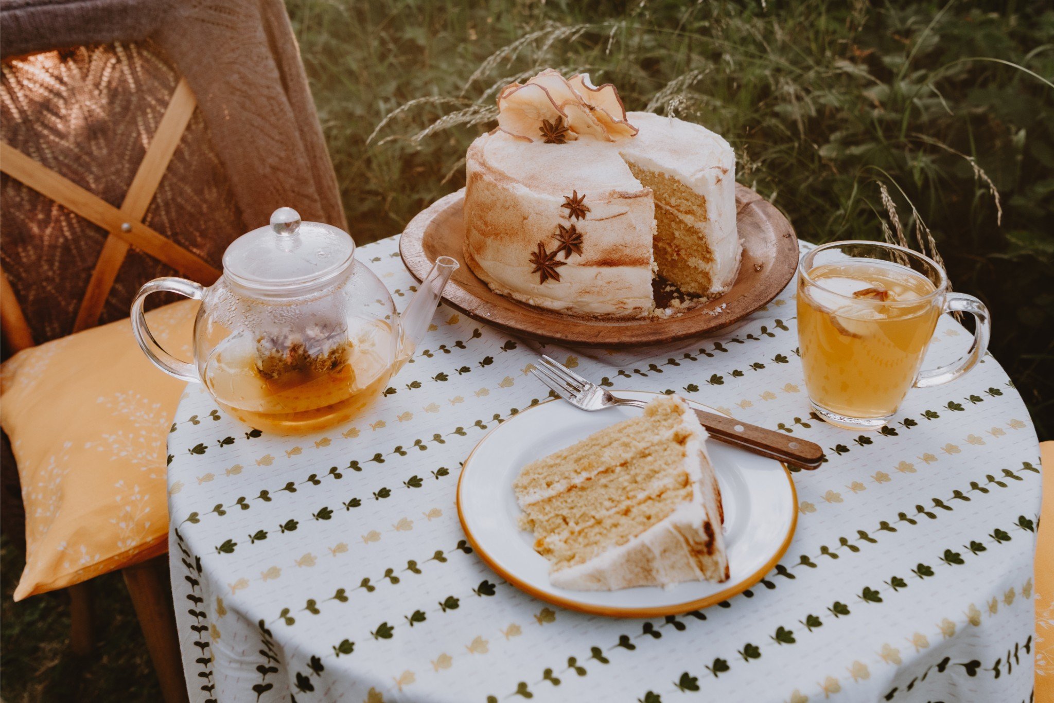 A slice of vanilla cake with white frosting on a plate, a whole cake on a wooden platter, a teapot with tea, a cup of tea, and a glass of apple juice are arranged on a table with a white tablecloth featuring green leaf patterns outdoors.