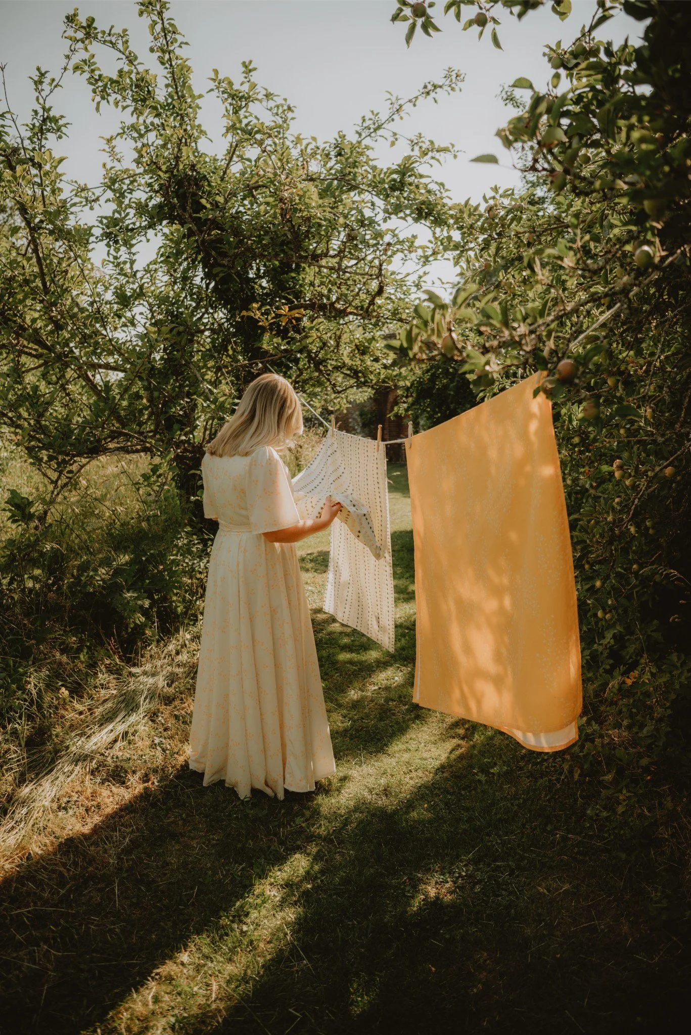 A woman in a long, light-colored dress hanging laundry on a clothesline in a lush, green garden with trees and grass, during daytime.